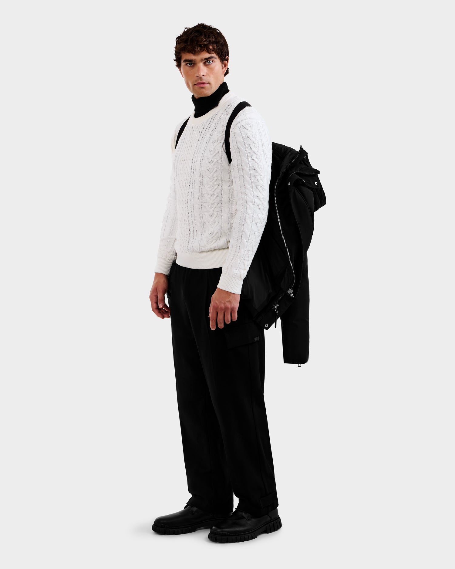 A man stands against a plain light background, wearing a white cable-knit sweater, black turtleneck, black pants, and black shoes while holding the Rudsak APOLLO - 6125693 Black coat over his shoulder.