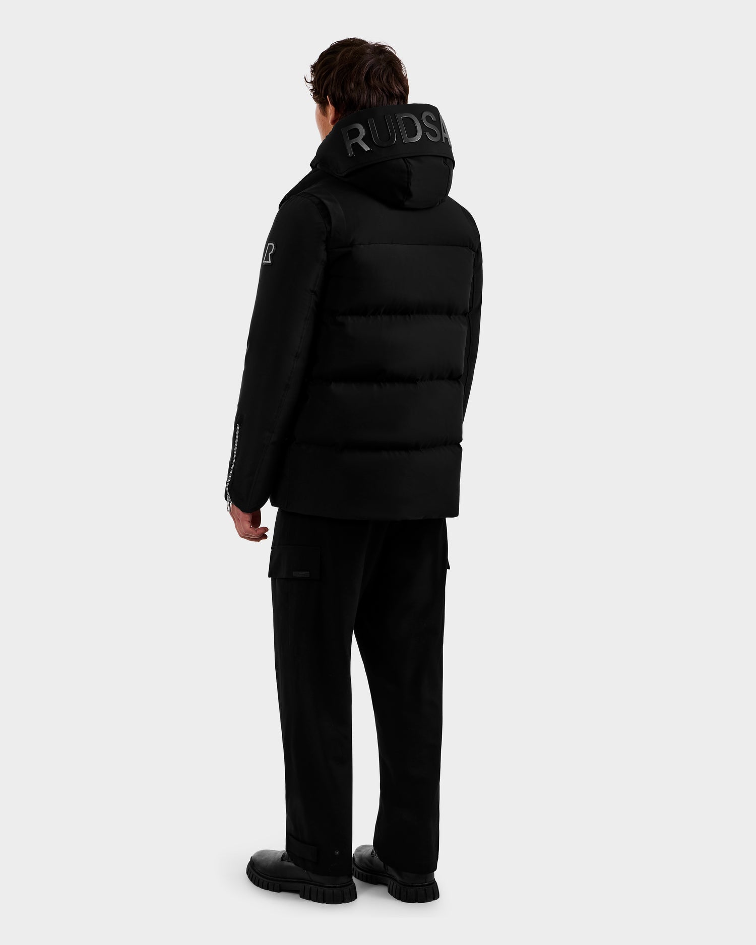 A person stands facing away wearing the Rudsak APOLLO - 6125693 Black puffer jacket with RUDSAK on the hood, black pants, and black shoes against a light gray background.
