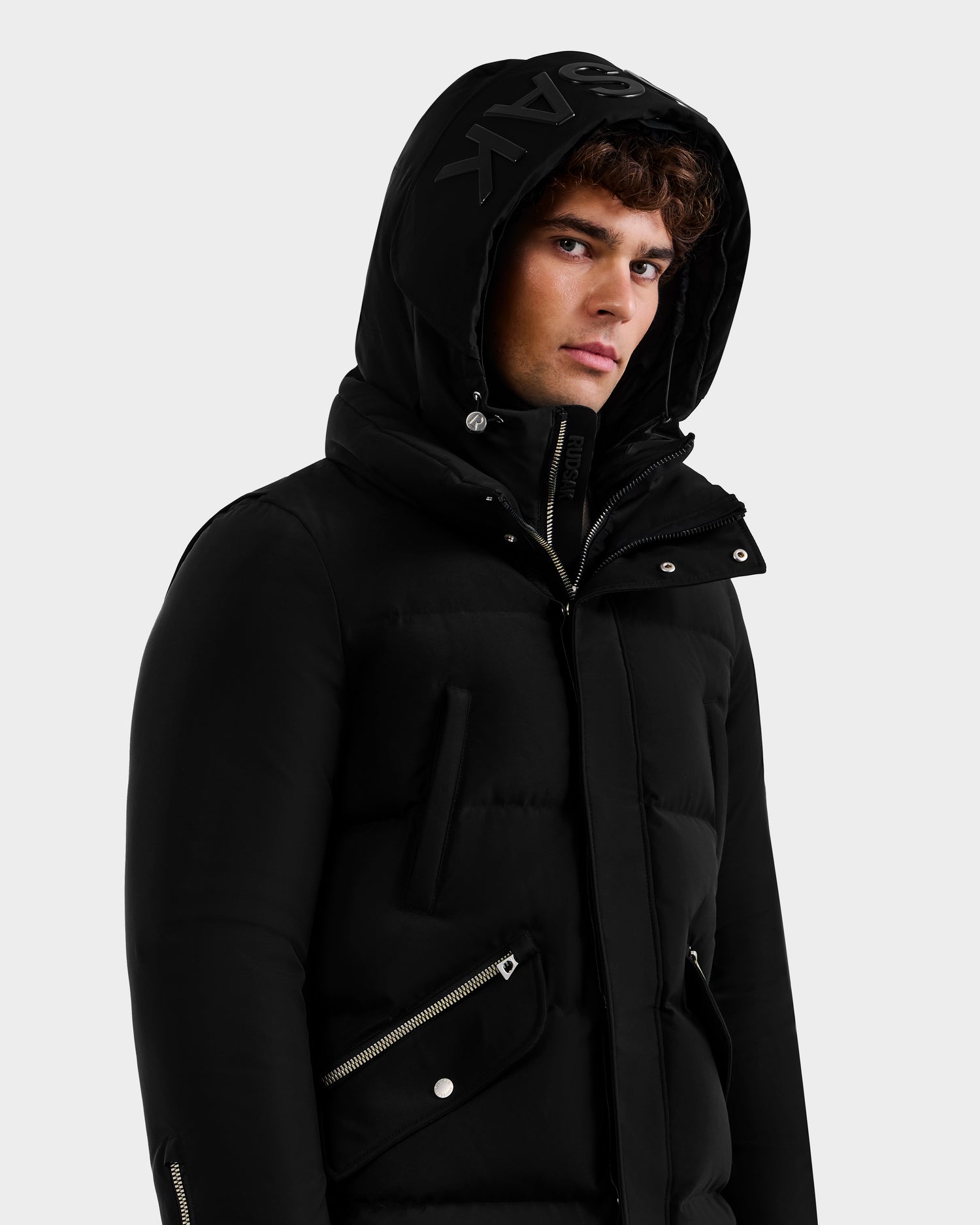 A young man wears the Rudsak APOLLO - 6125693 Black hooded puffer jacket with zippered pockets, a raised collar, and bold black lettering on the hood, looking slightly to the side against a plain light background.
