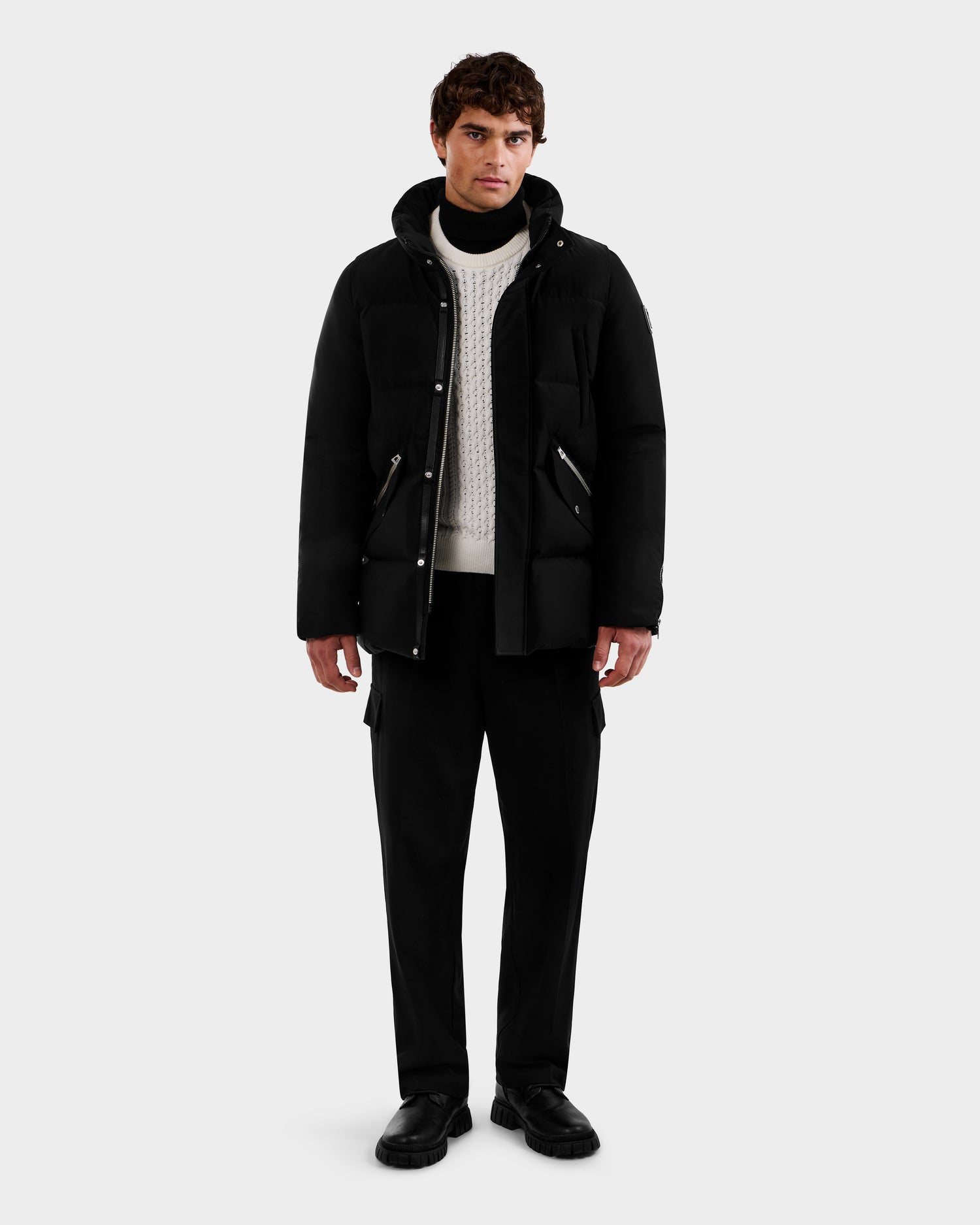 A man faces forward in a Rudsak APOLLO - 6125693 black puffer jacket, layered over a white knit sweater with black pants and shoes, standing against a plain light gray background.