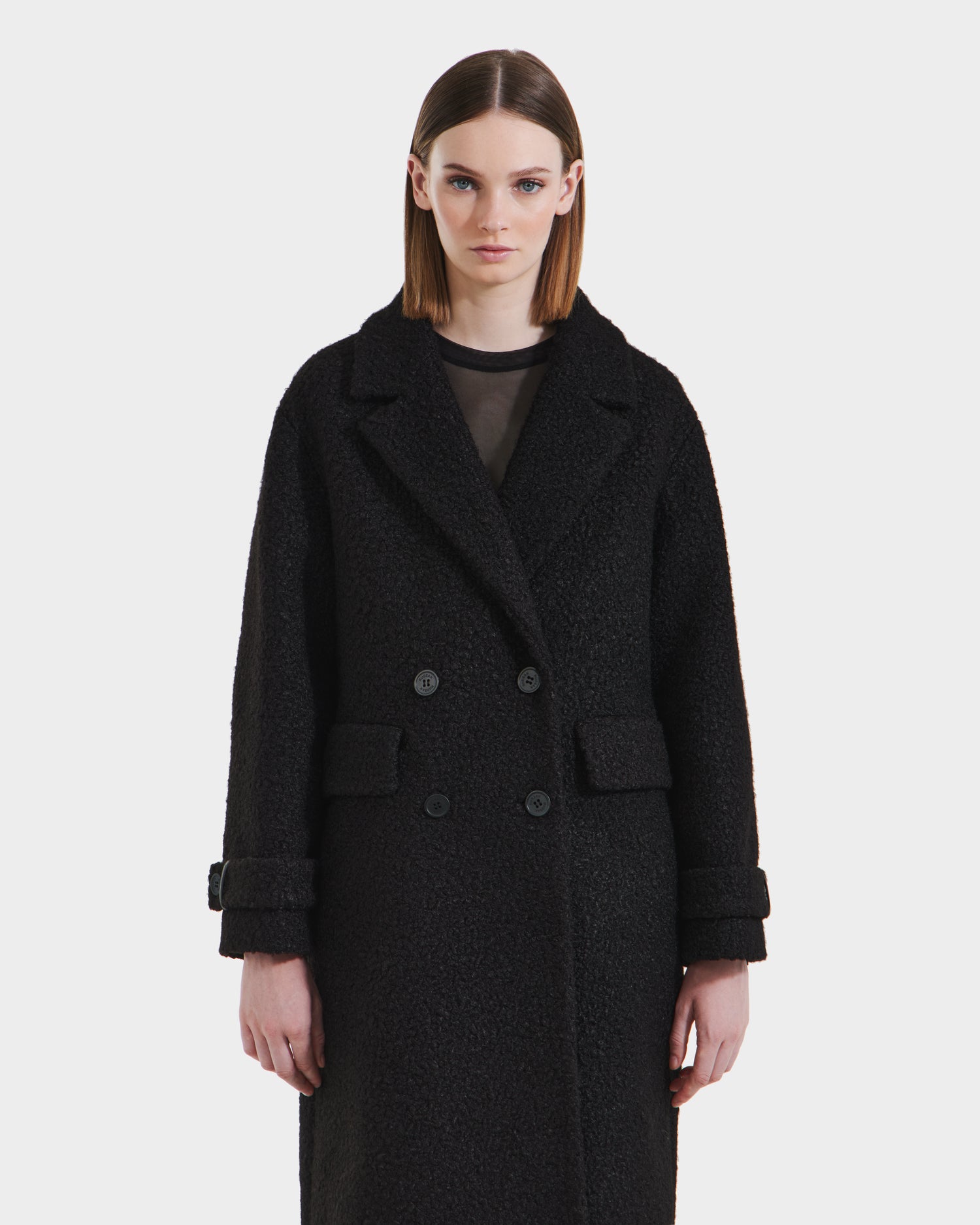 A woman with straight brown hair models the Rudsak ARABELLA - 8125621 Black overcoat, featuring large lapels and cuffed sleeves, standing against a plain white background.