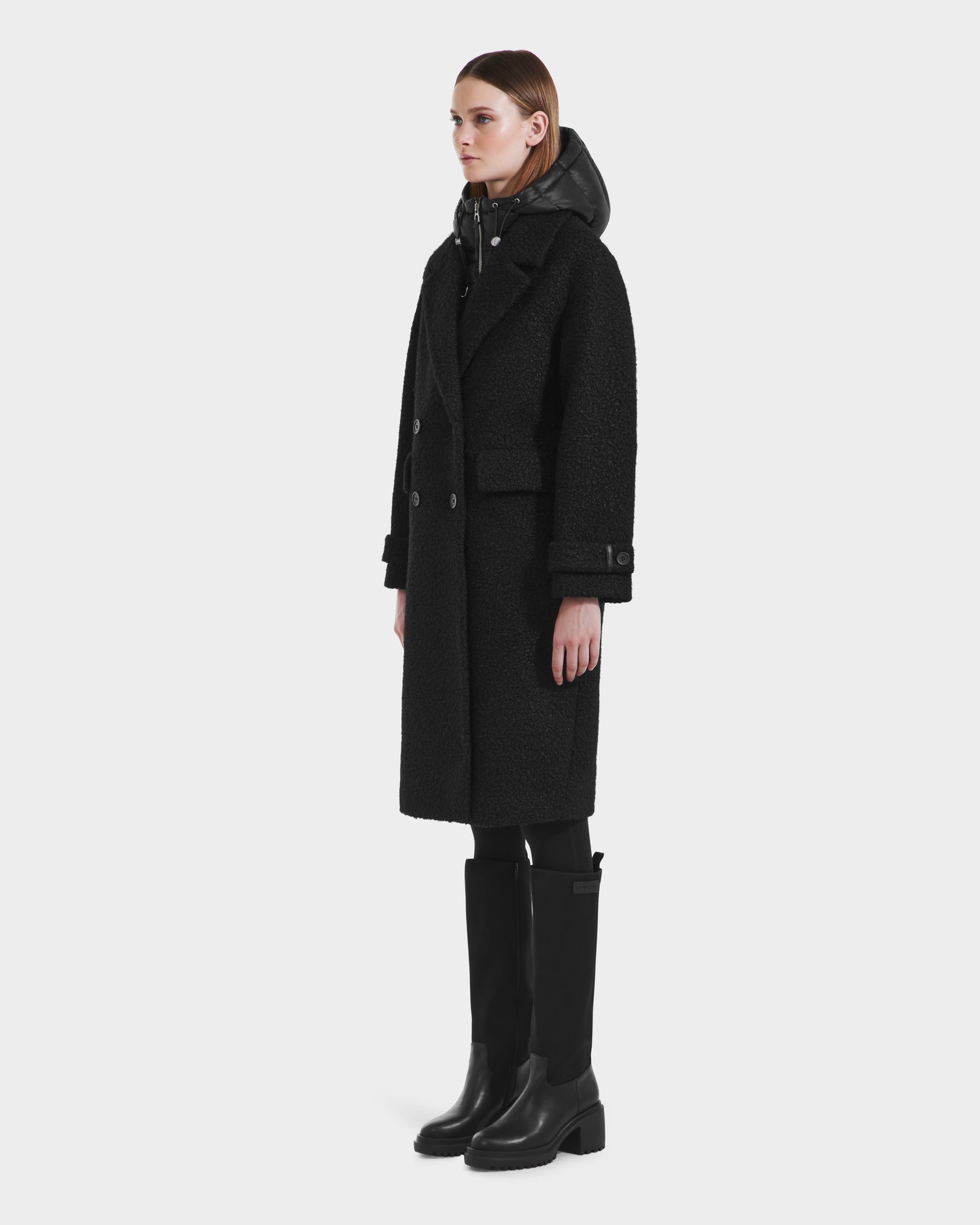 A woman stands facing left in a long black ARABELLA - 8125621 coat by Rudsak, layered over a black hooded jacket, black leggings, and knee-high boots against a plain white background.