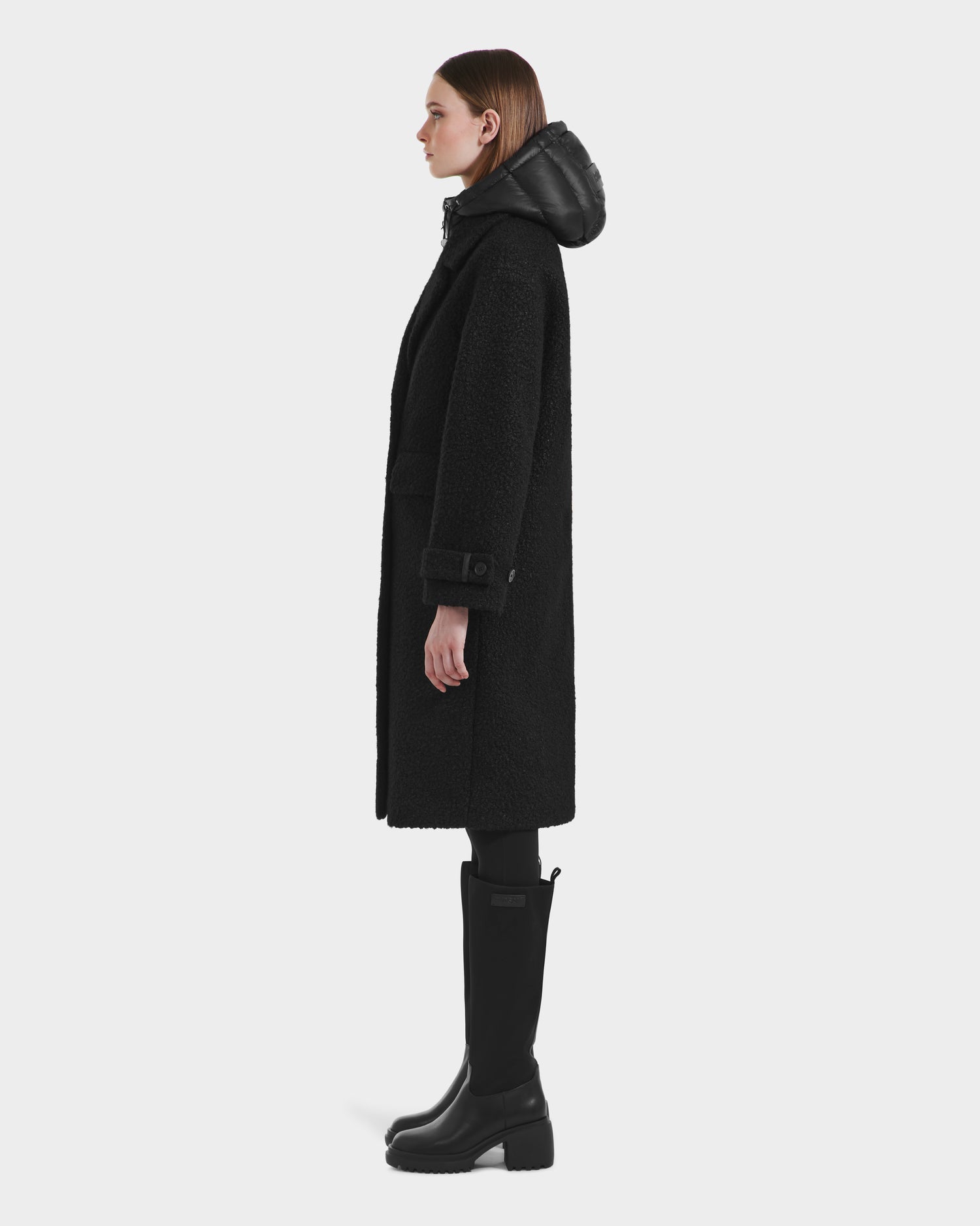 A woman stands in profile wearing the Rudsak ARABELLA - 8125621 Black, a long textured hooded coat, paired with black leggings and knee-high boots, set against a plain light background.