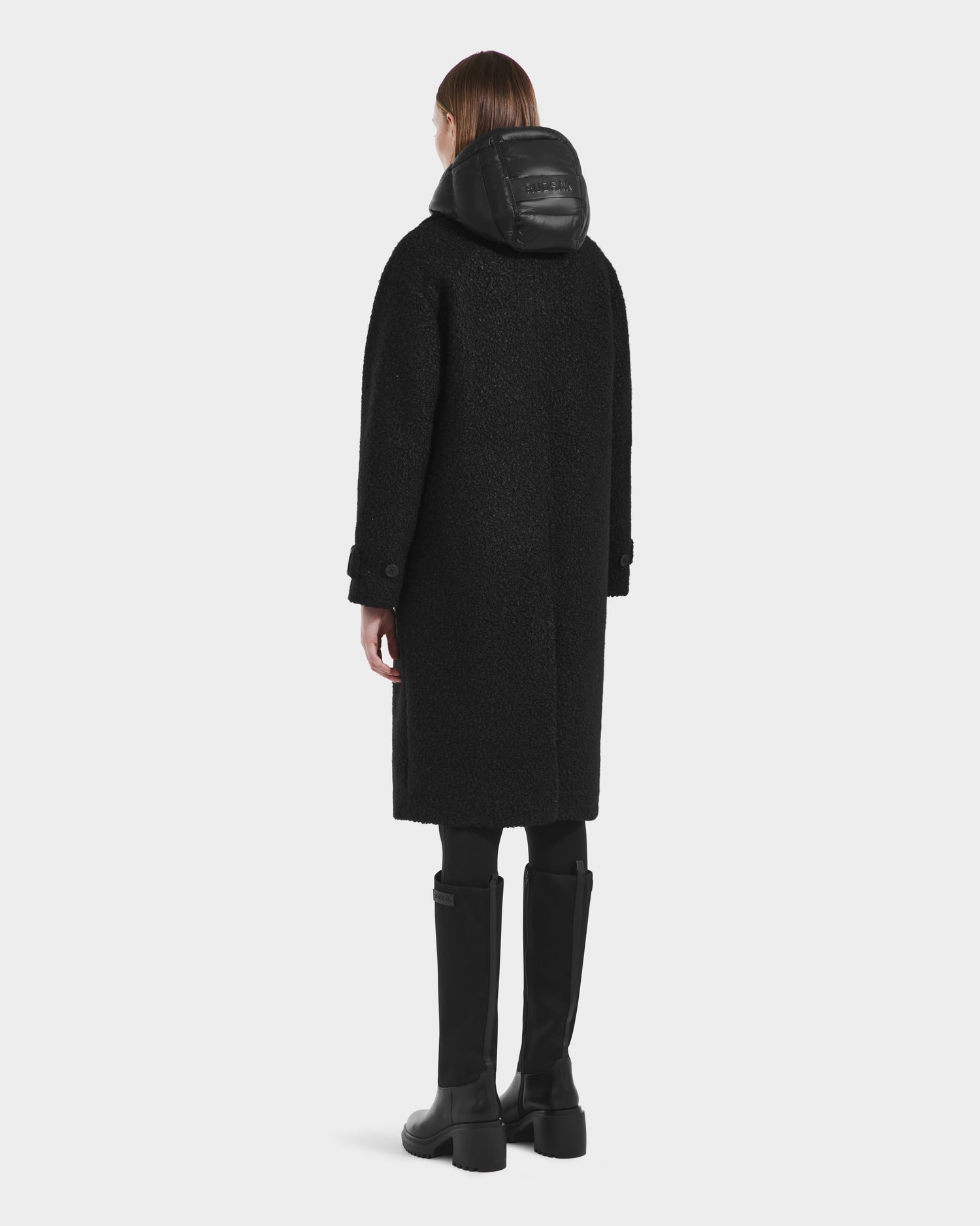 A person with straight brown hair, wearing the Rudsak ARABELLA - 8125621 Black women’s overcoat, black pants, tall black boots, and a shiny hood, stands facing away from the camera.