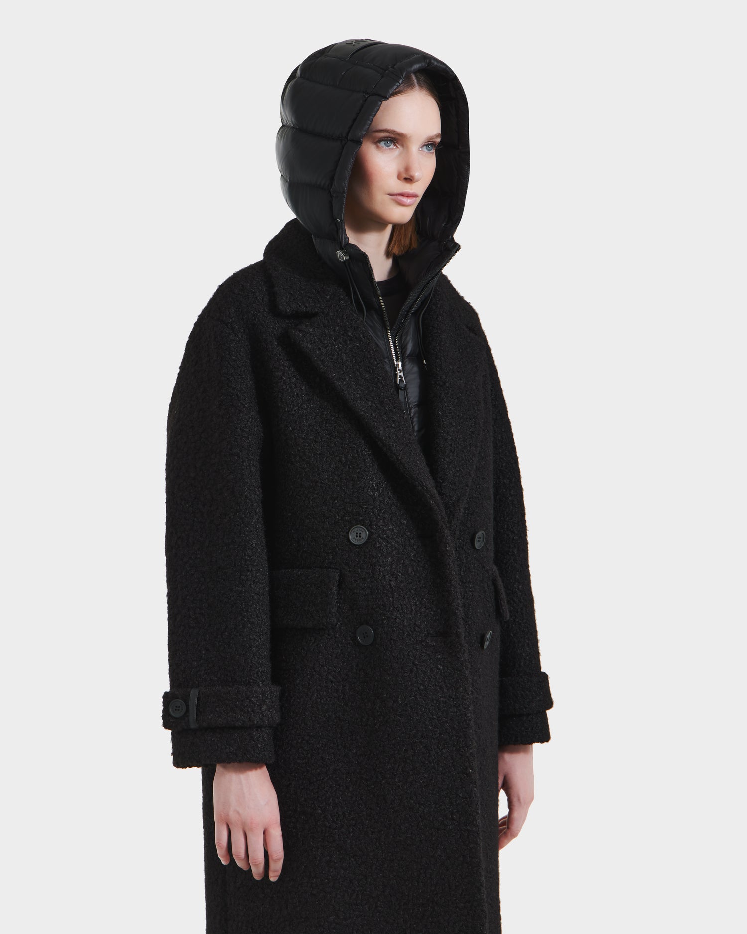 A woman models the Rudsak ARABELLA - 8125621 Black textured double-breasted overcoat with buttoned cuffs and a shiny black puffer hood, standing slightly to the right against a white background—stylish, versatile outerwear for any occasion.