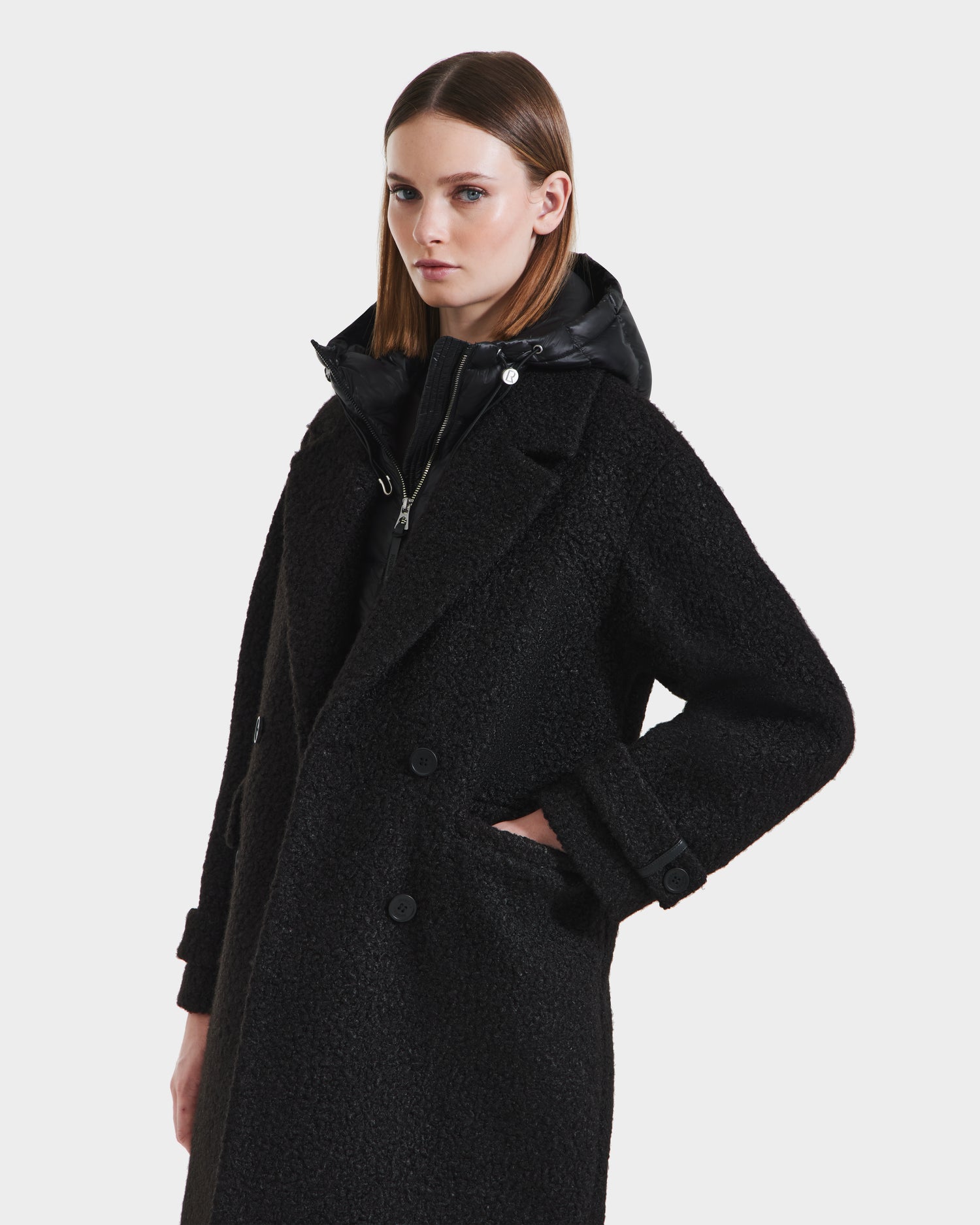 A woman with straight brown hair wears the Rudsak ARABELLA - 8125621 Black textured, double-breasted women’s overcoat with a black zip-up inner layer, standing with one hand in her pocket against a plain light background.