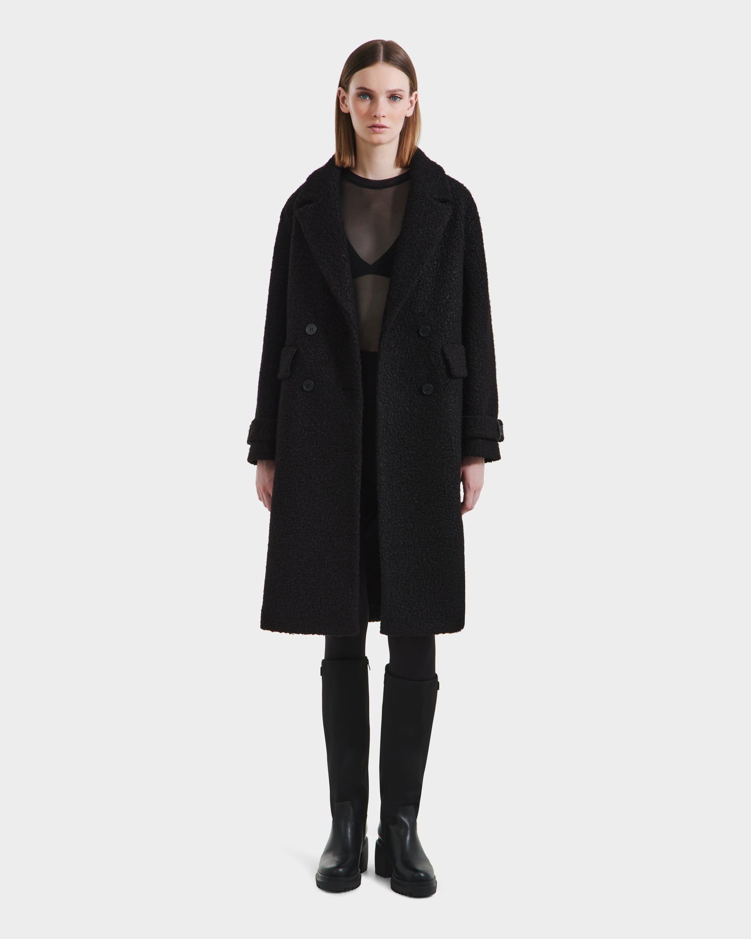 A woman faces forward against a plain white background, wearing the Rudsak ARABELLA - 8125621 Black textured overcoat over a sheer black top, black shorts or skirt, leggings, and knee-high boots.
