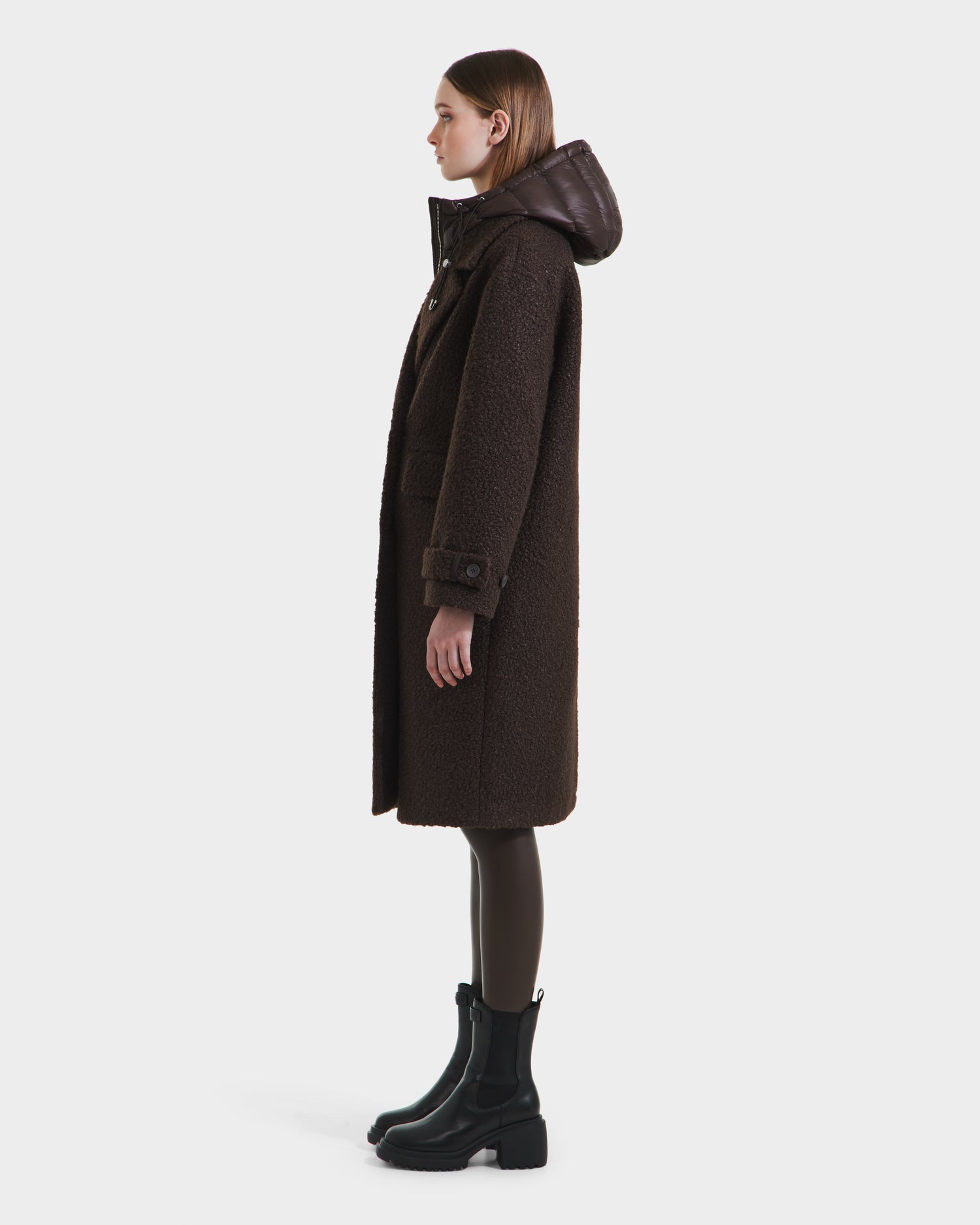A woman in profile wears the ARABELLA - 8125621 Espresso long, dark brown women’s overcoat by Rudsak, featuring a shiny hood. She pairs it with dark leggings and black ankle boots against a plain white background.