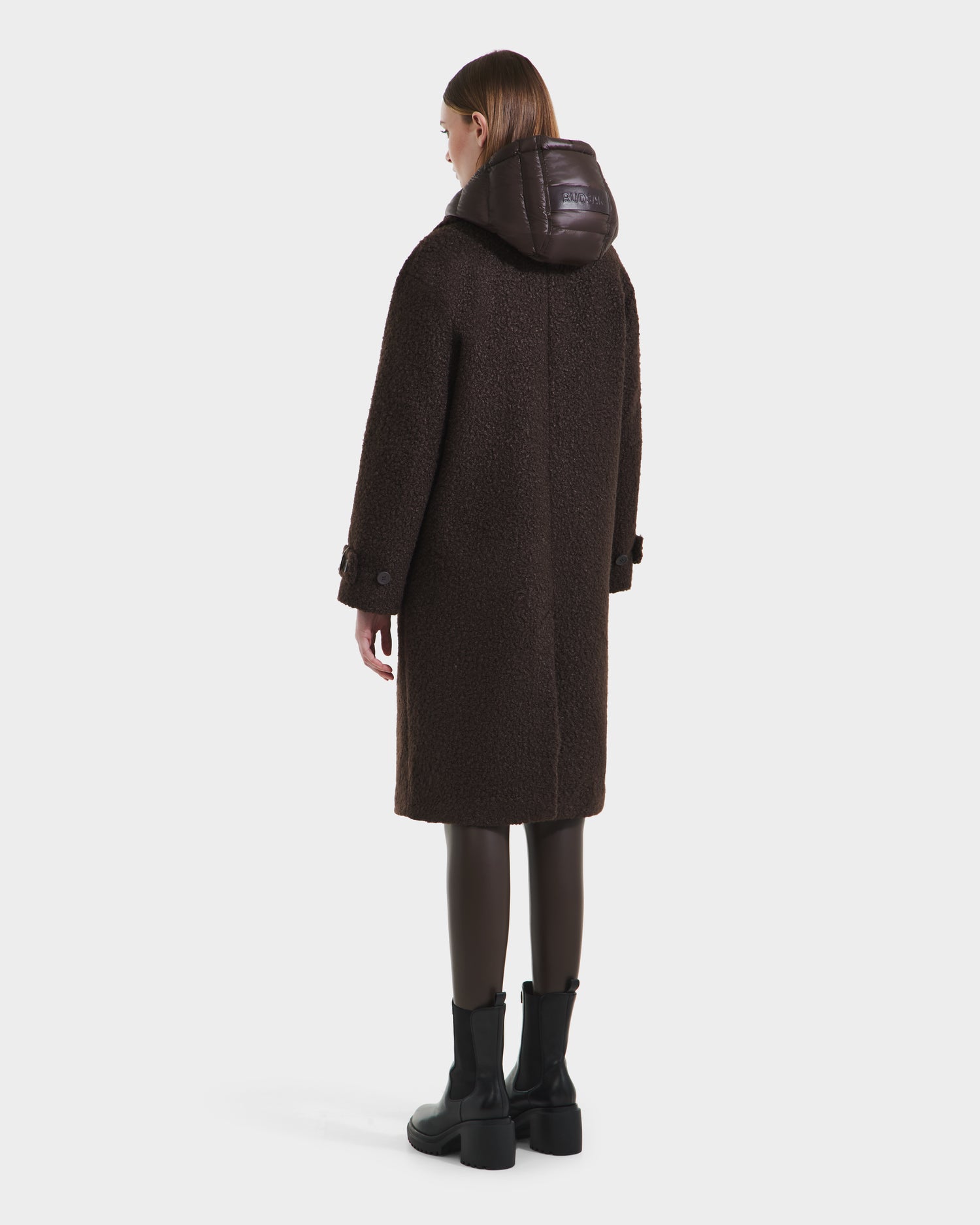 A woman faces away, wearing the Rudsak ARABELLA - 8125621 Espresso long dark brown textured overcoat with a shiny hood, paired with black leggings and boots, set against a plain light background.