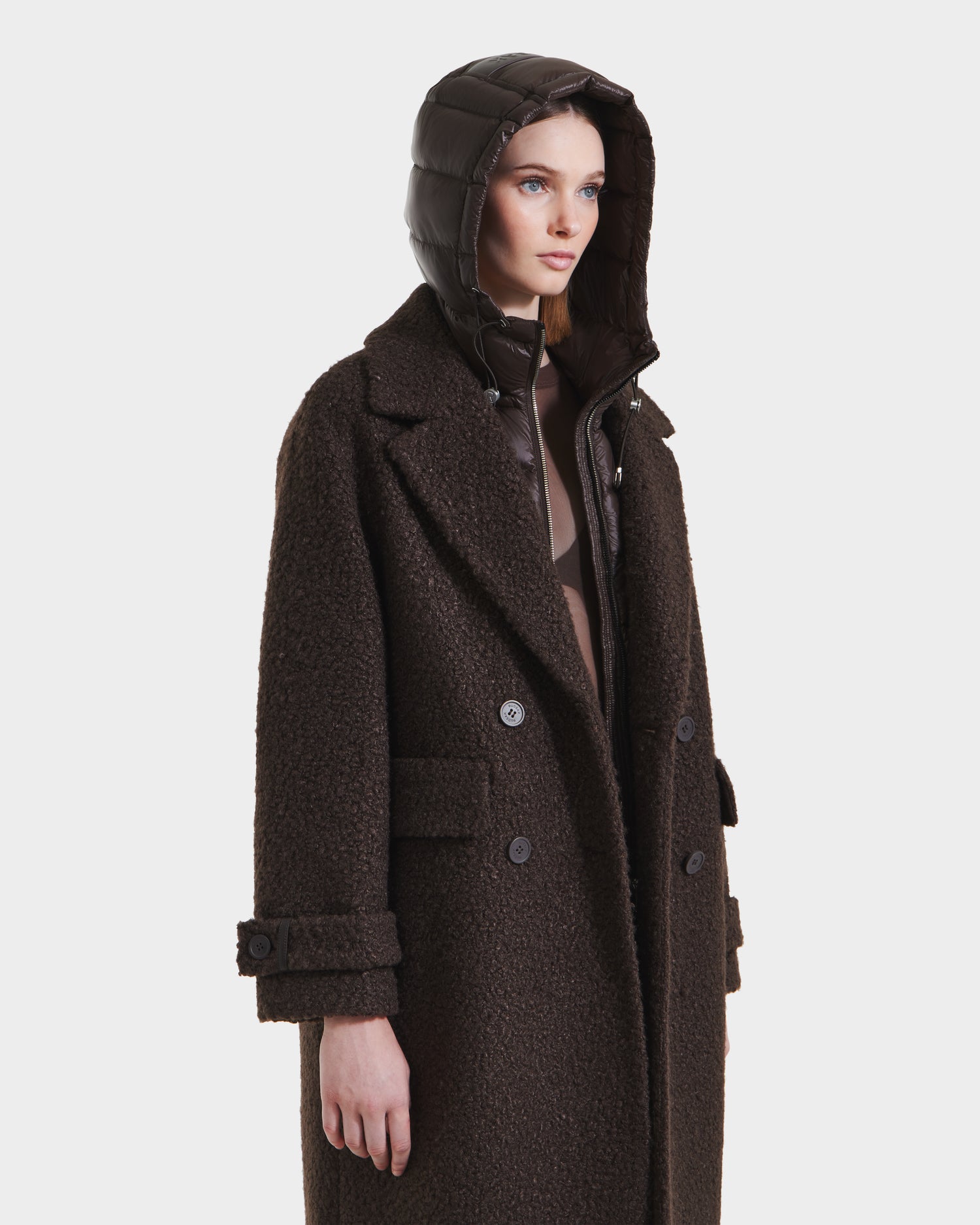 A woman in the ARABELLA - 8125621 Espresso overcoat by Rudsak, featuring a dark brown textured finish, button details, and a padded hood, stands against a plain white background. This outerwear piece offers both style and comfort.