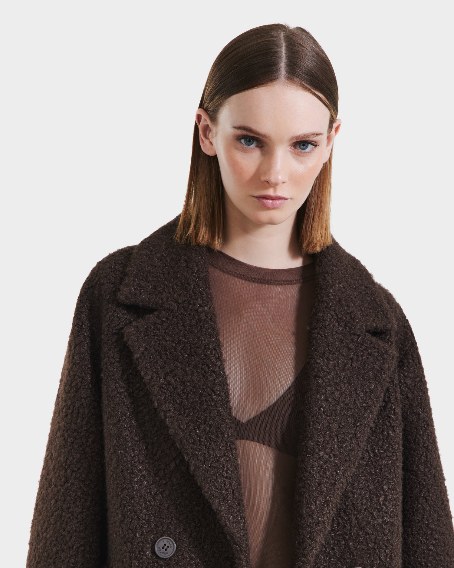 A woman with straight, shoulder-length brown hair models the Rudsak ARABELLA - 8125621 Espresso, a textured dark brown womens overcoat, layered over a sheer brown top and bralette, against a plain white background.