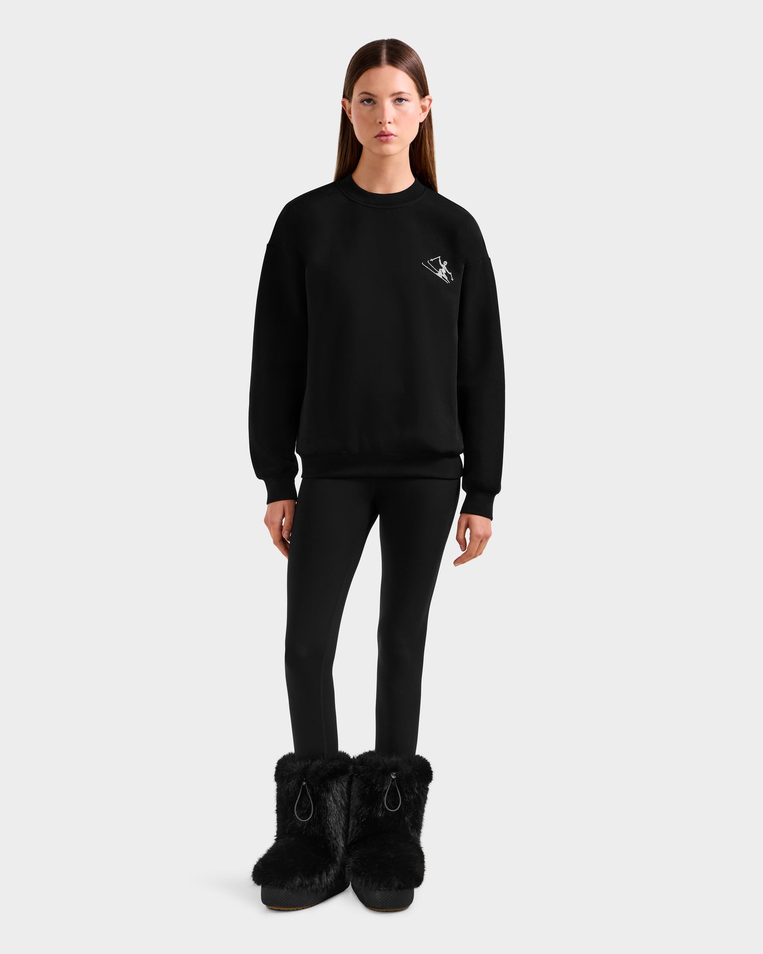 A woman with straight brown hair and a neutral expression wears the Rudsak ARIA - 8525661 Black women’s crewneck sweatshirt with a small white ski graphic, paired with black leggings and fluffy black boots, against a plain background.