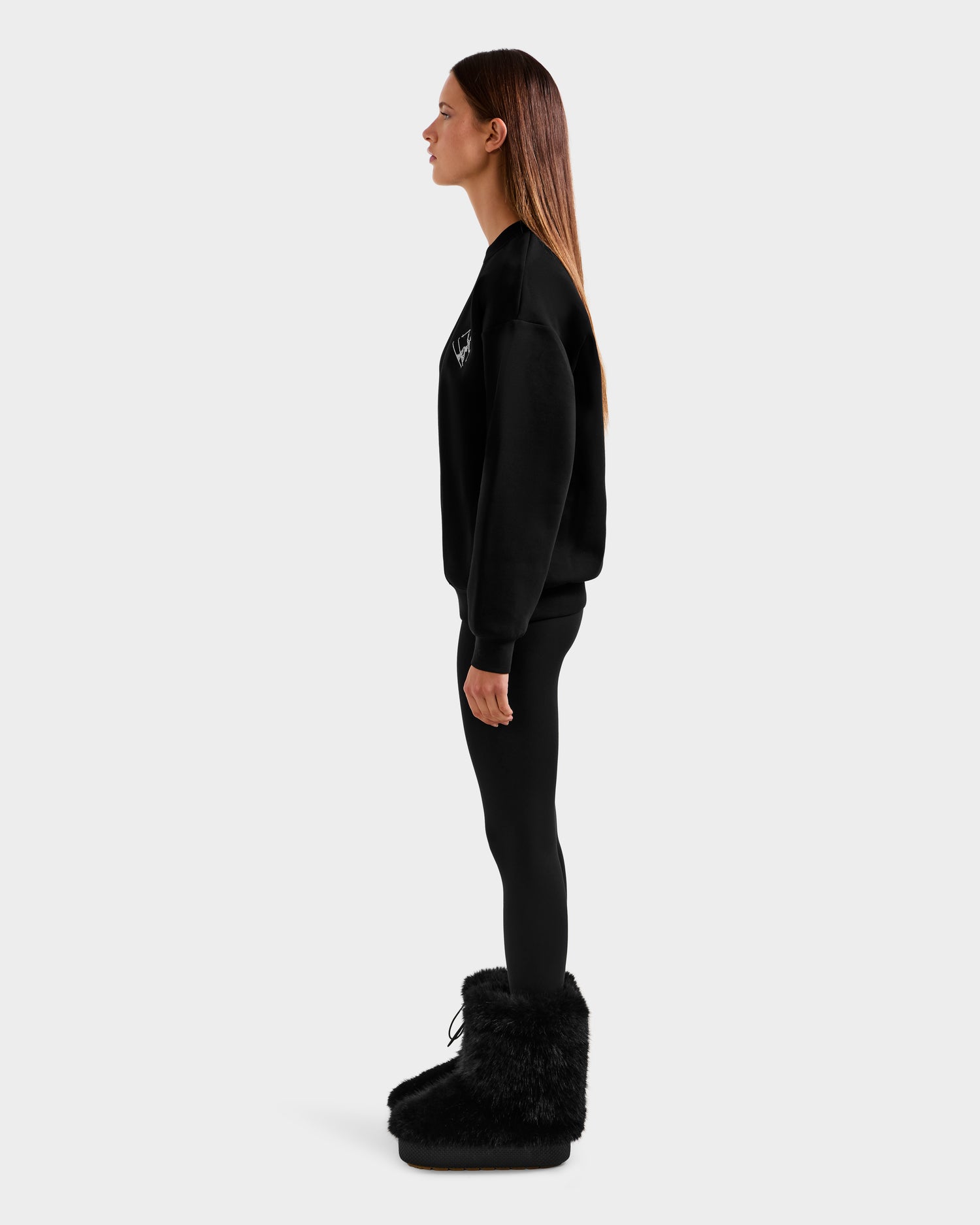 A woman stands in profile against a plain background, wearing Rudsak’s ARIA - 8525661 Black crewneck sweatshirt and leggings with large black furry boots. Her straight brown hair is down, and her arms rest at her sides.