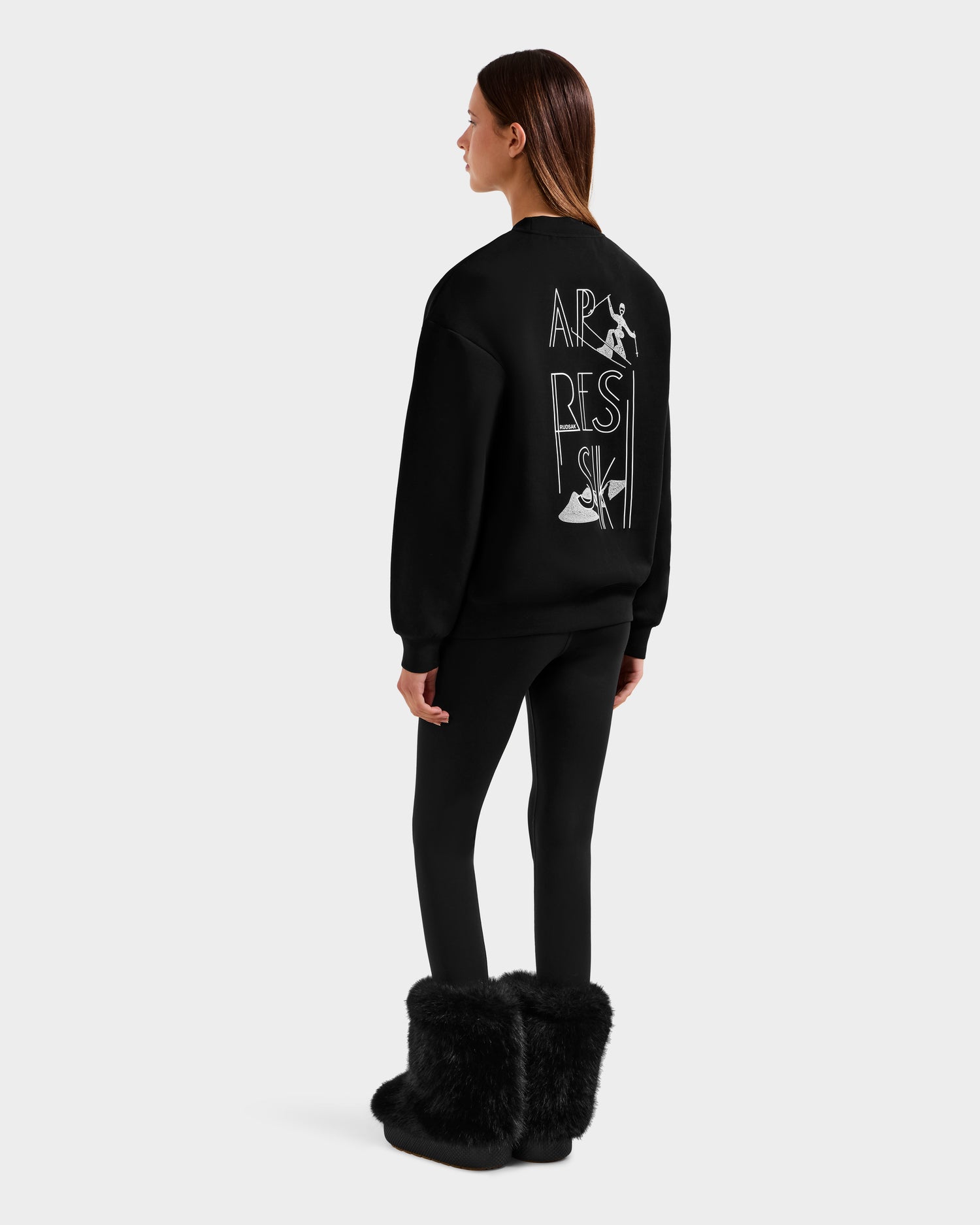A woman faces away, wearing the Rudsak ARIA - 8525661 Black women's crewneck sweatshirt with white abstract text and graphics on the back, paired with black leggings and furry boots—an ideal winter loungewear look.