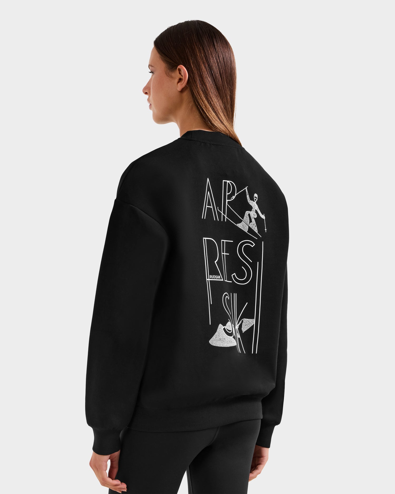 A woman with straight brown hair, facing away to the left, wears the Rudsak ARIA - 8525661 Black women’s crewneck sweatshirt featuring white line art, a ski graphic with “APRES SKI” on the back, and small skier and ski boots drawings.
