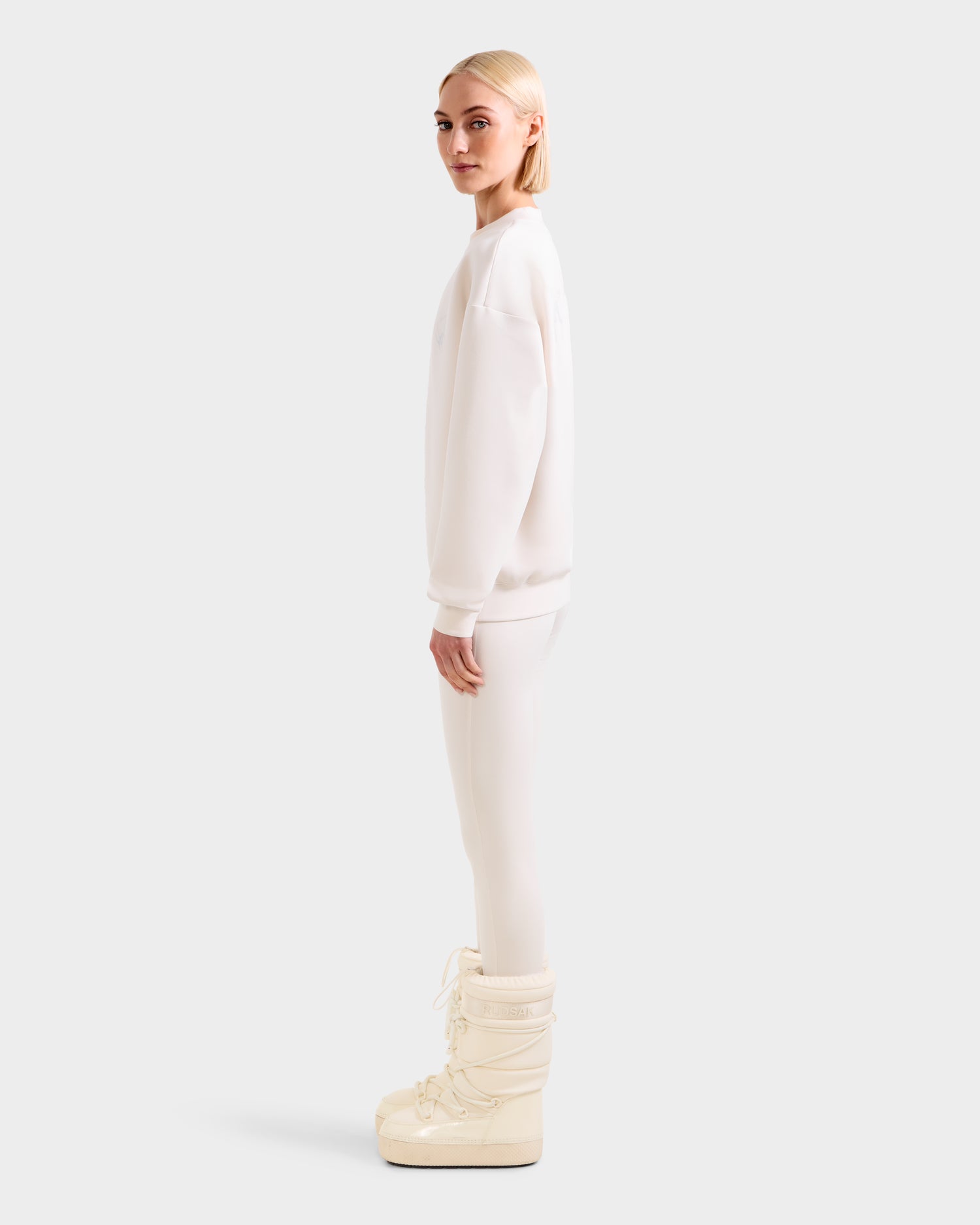 A blonde-haired person in profile wears the Rudsak ARIA - 8525661 Vanilla women’s crewneck sweatshirt with matching leggings and large white boots, set against a plain light gray background.