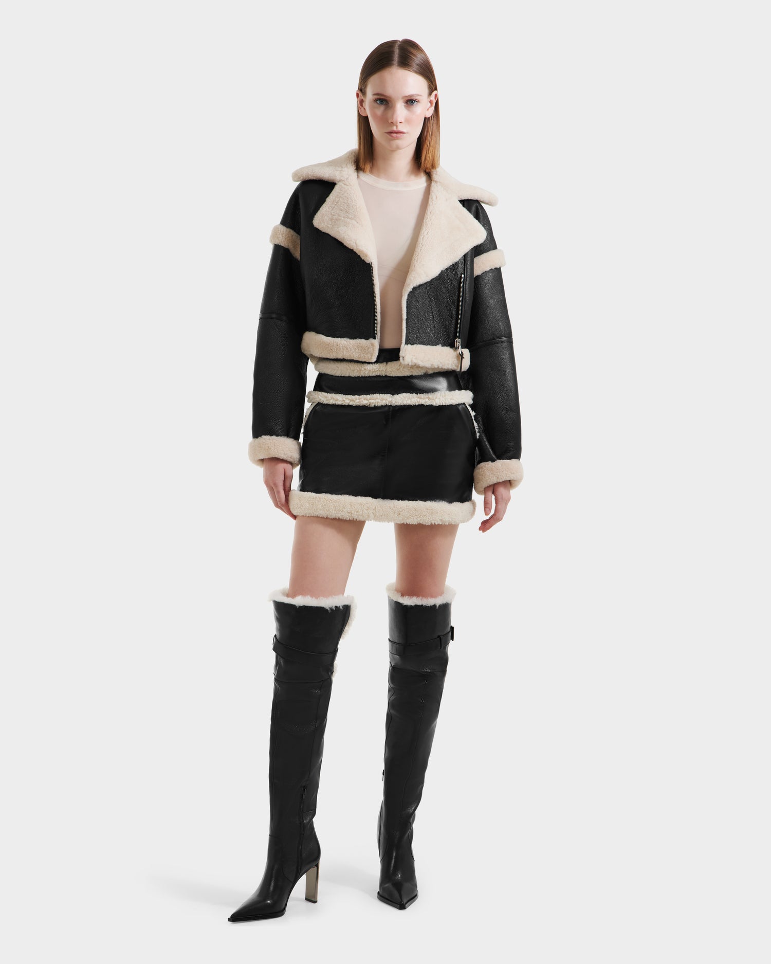 A woman faces forward against a white background, wearing the Rudsak ARIANNA - 8125504 Black knee-high heeled boots, styled with a luxe shearling cropped jacket, matching mini skirt, and light top.