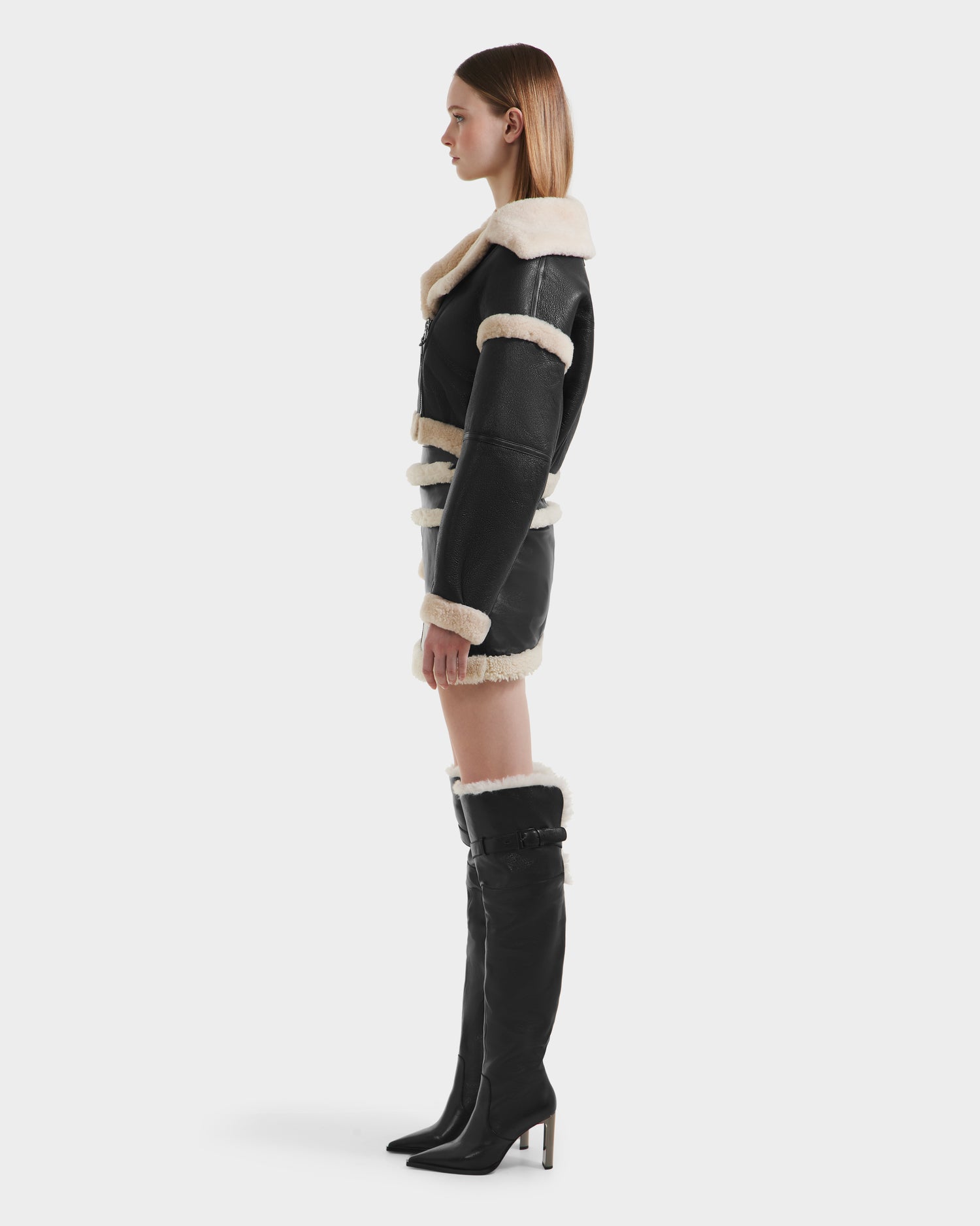A woman in profile wears the Rudsak ARIANNA - 8125504 Black luxe shearling cropped jacket with white trim, a matching mini skirt, and tall black boots topped with white shearling, set against a plain white background.