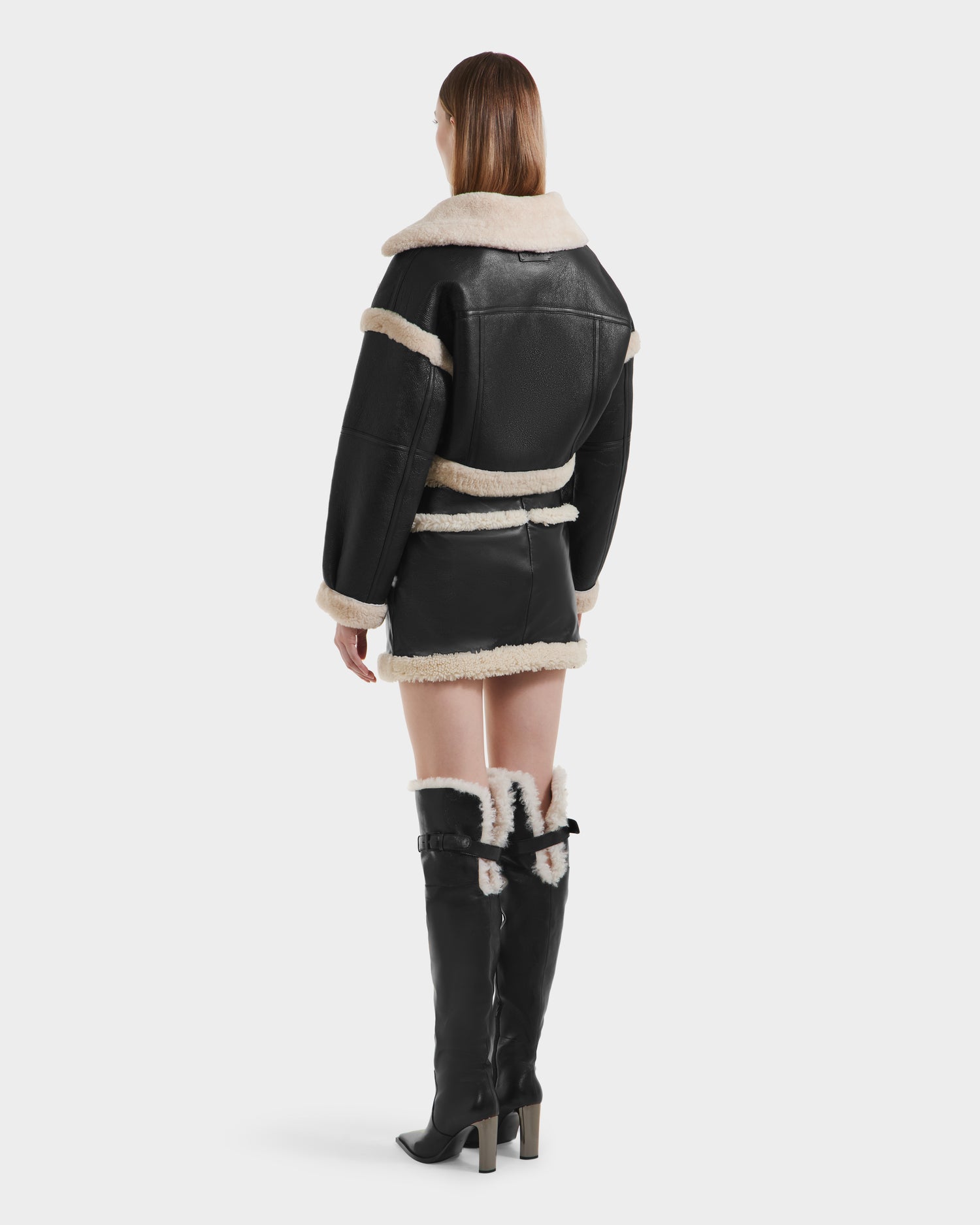 A person stands facing away, wearing the Rudsak ARIANNA - 8125504 Black tall boots with fur trim, paired with a luxe shearling mini skirt and matching cropped jacket. Their straight, shoulder-length hair contrasts the plain white background.