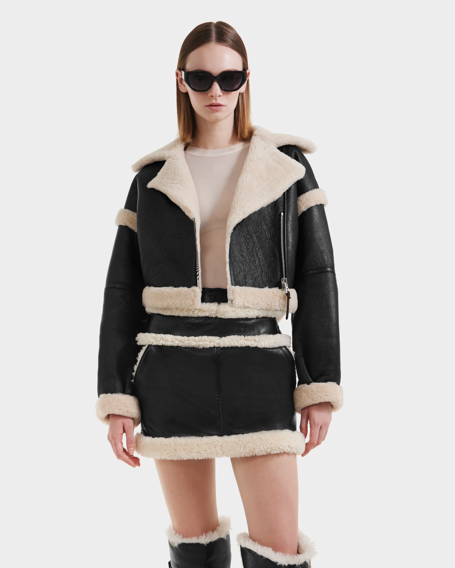 A woman wearing Rudsak’s ARIANNA - 8125504 Black cropped shearling jacket with leather trim, a matching mini skirt with cream fur, black sunglasses, a beige top, and knee-high black boots stands against a white background.