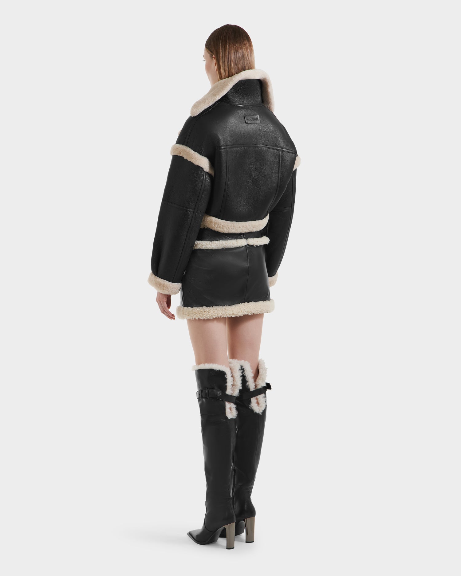 A woman faces away, wearing the Rudsak ARIANNA - 8125504 Black cropped shearling jacket and mini skirt set, styled with black knee-high boots featuring cream shearling trim and contrasting light-colored accents.