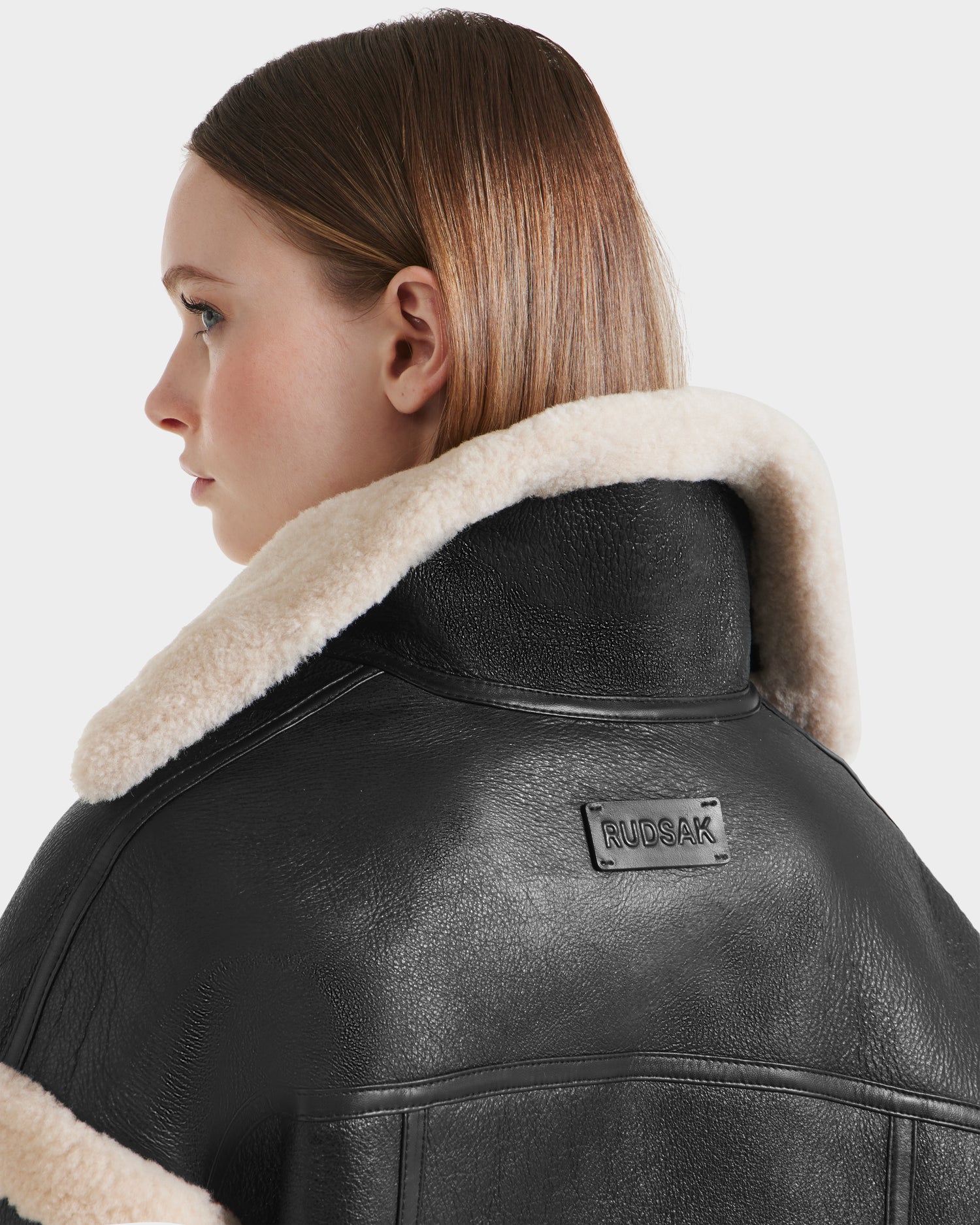 A woman with straight, light brown hair looks to the side, wearing the Rudsak ARIANNA - 8125504 Black cropped shearling jacket with a cream shearling collar. A modern minimalist look, it features a logo on the back. The background is plain white.