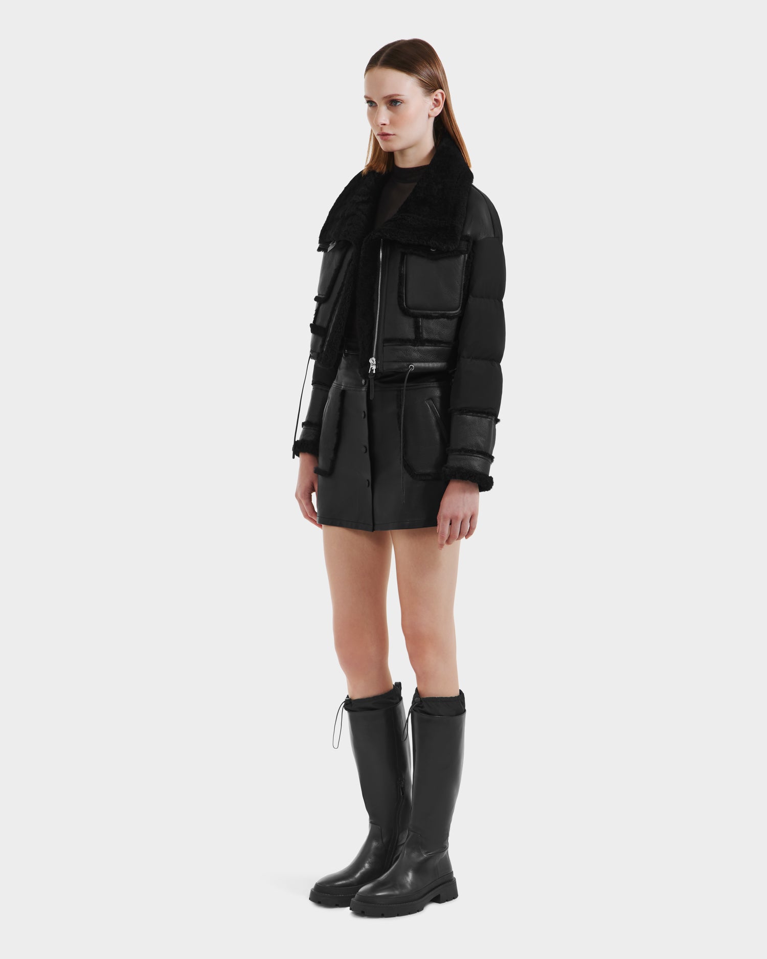 A woman with straight, shoulder-length hair and a neutral expression wears the Rudsak ARIELLA - 8125505 Black women’s cropped shearling puffer against a plain background, paired with a black mini skirt and knee-high black boots.