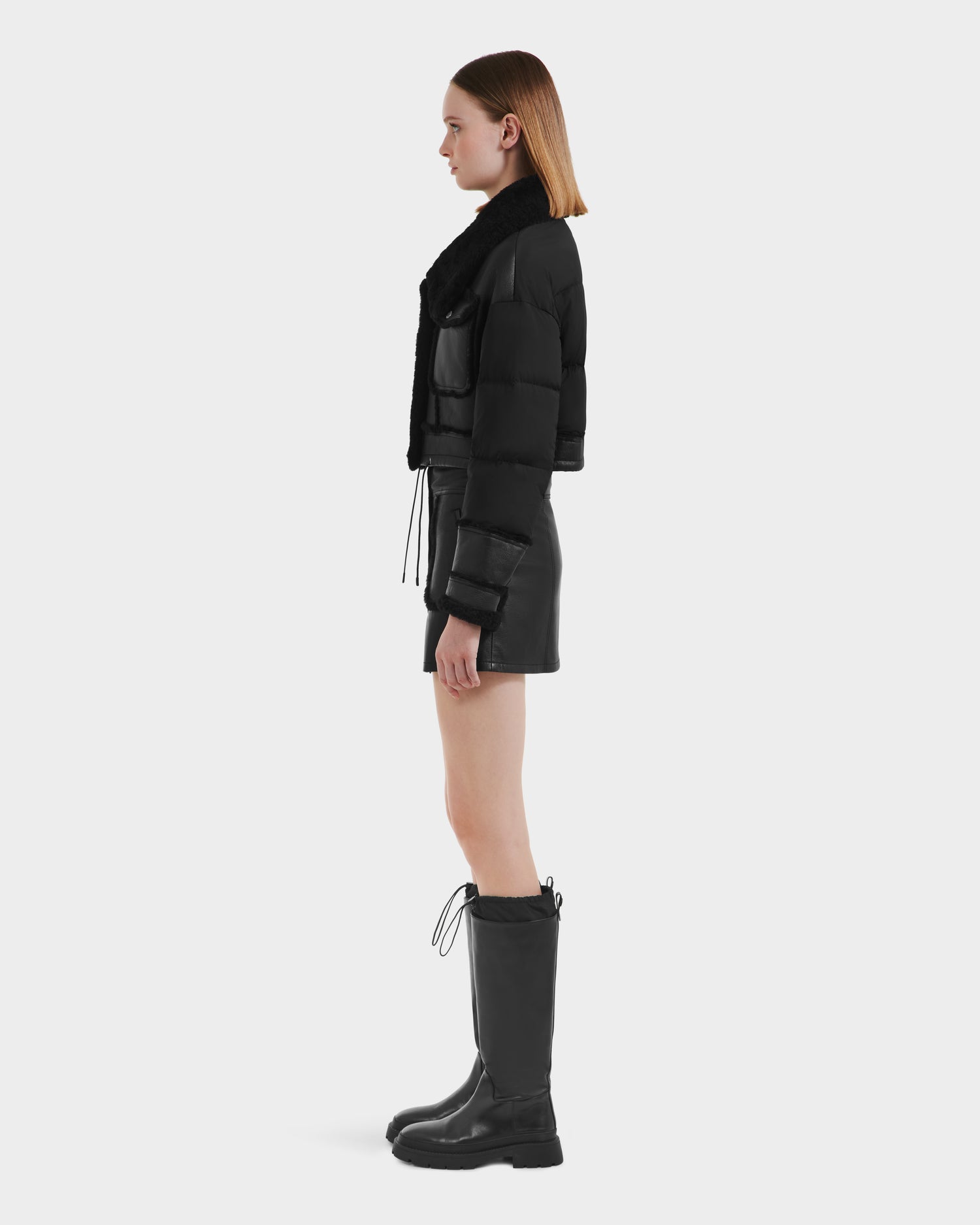 A woman stands in profile wearing the Rudsak ARIELLA - 8125505 Black cropped shearling puffer jacket, styled with a black mini skirt and tall black boots—an effortlessly chic cold-weather look set against a plain white background.