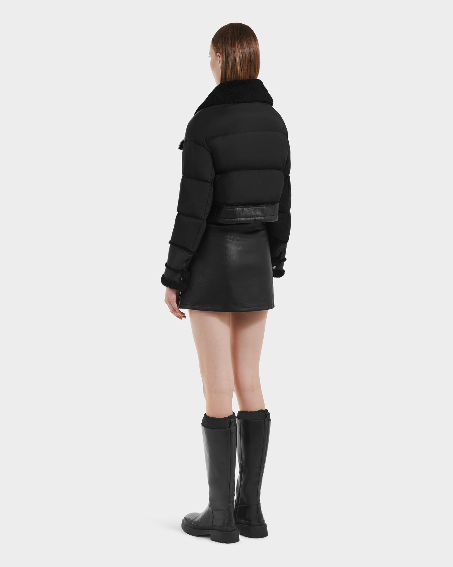 A woman faces away, modeling Rudsaks ARIELLA - 8125505 Black cropped shearling puffer with a fur collar, styled with a black leather mini skirt and knee-high boots against a plain white background.