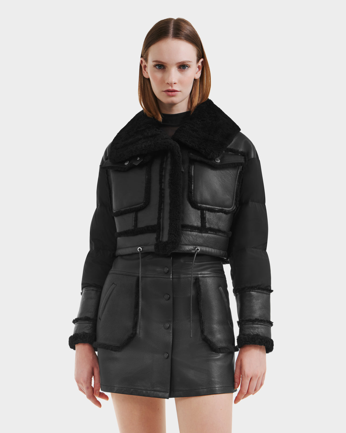 A woman with straight brown hair models the Rudsak ARIELLA - 8125505 Black cold-weather jacket with shearling trim and a matching black leather mini skirt, standing against a plain white background.