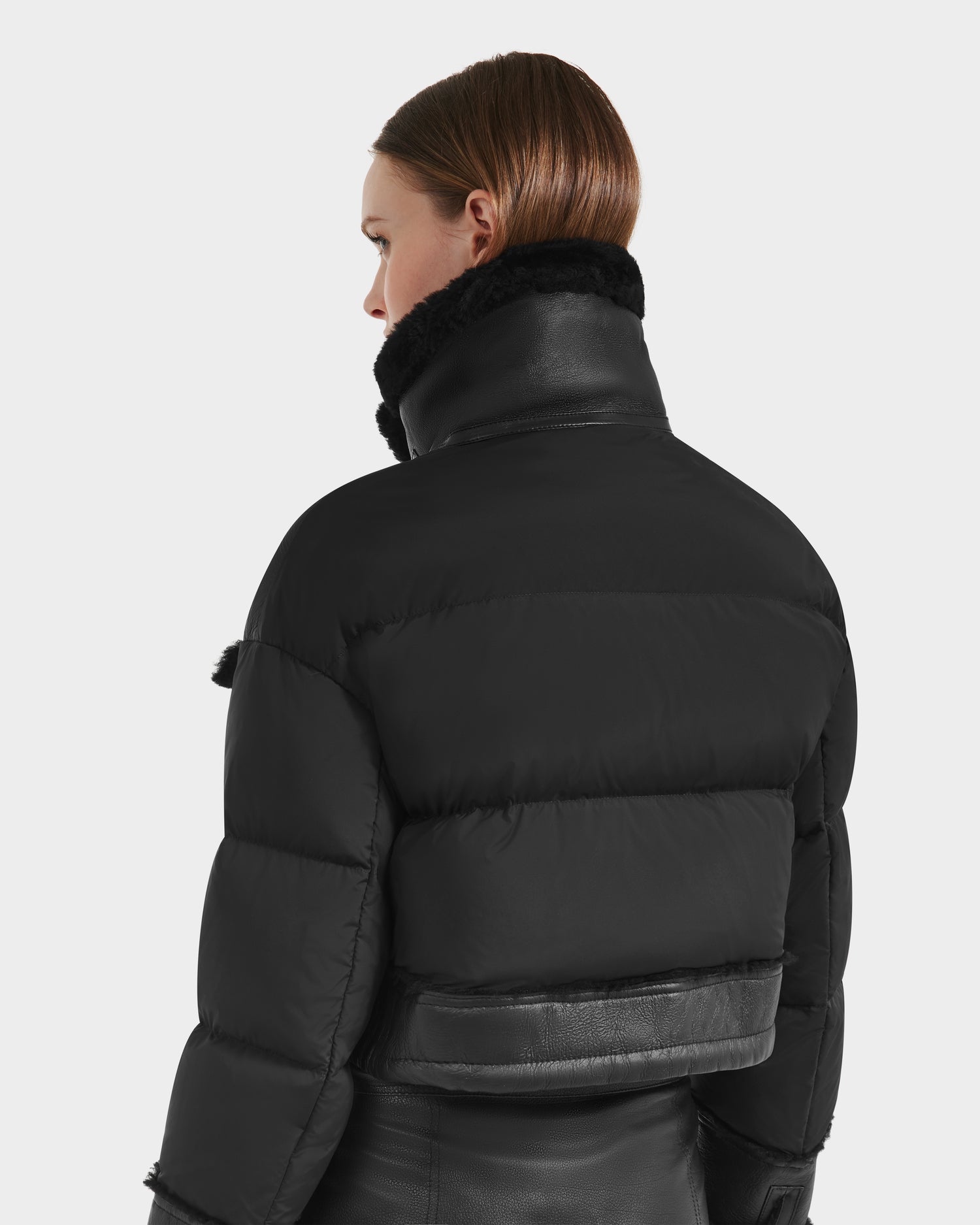 A person with straight, shoulder-length brown hair is seen from the back wearing the Rudsak ARIELLA - 8125505 Black puffer jacket, featuring a high textured collar and leather accents—perfect for cold weather—against a plain light background.