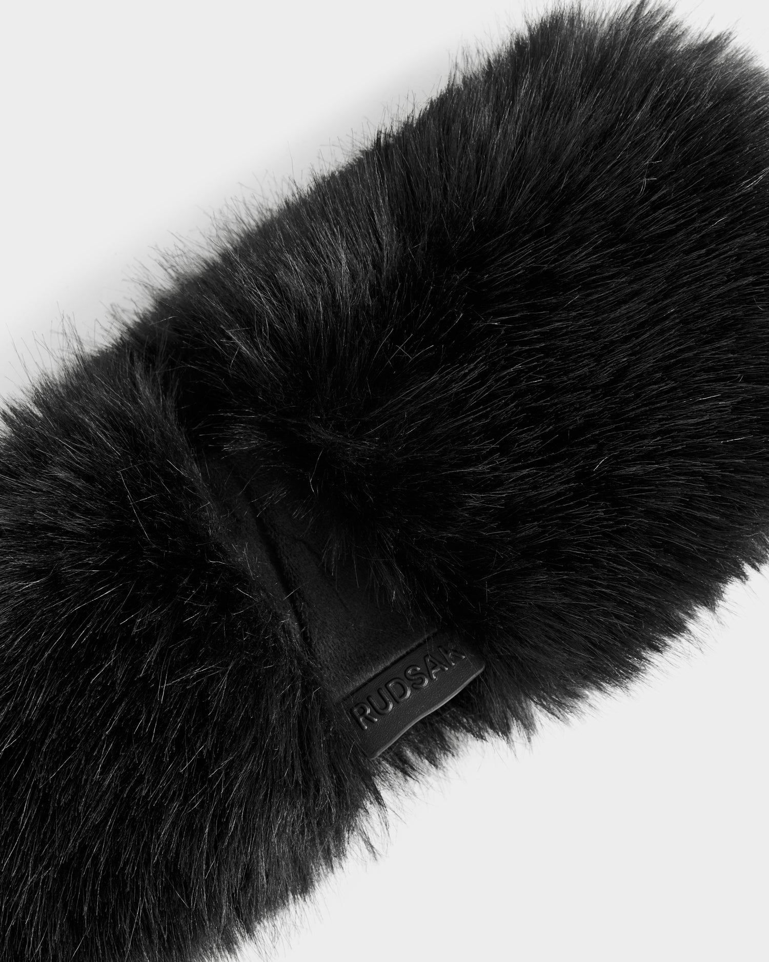 Close-up of the ARLETTE - 8425544 Black women's faux fur headband by Rudsak, featuring a chic leather tag embossed with the brand name. Set against a white background, it's the perfect accessory to elevate your winter fashion.
