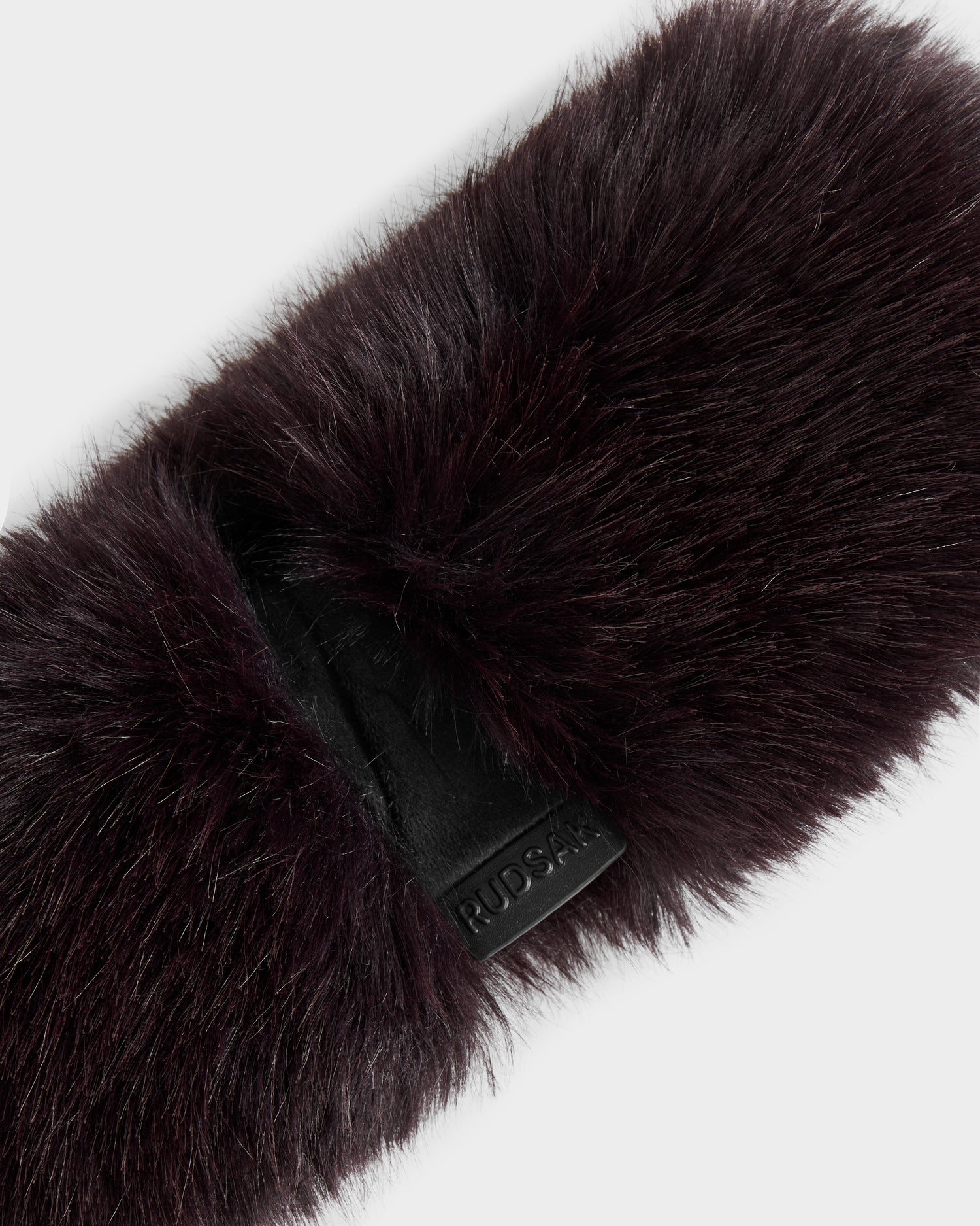 Close-up of the ARLETTE - 8425544 Bordeaux women’s faux fur headband by Rudsak, featuring a black leather tag embossed with the brand name, set against a plain white background—a cozy winter accessory.