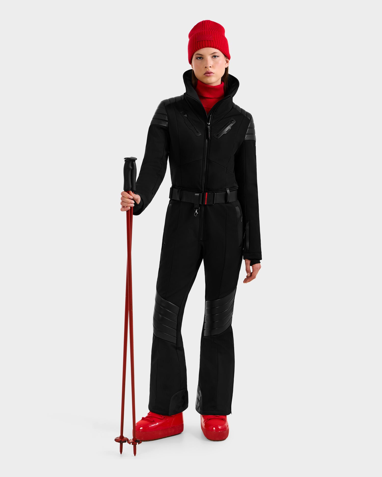 A person wears the Rudsak ARLEY - 8124717 Black fitted one-piece ski suit, paired with a red beanie, red ski boots, and holding red ski poles against a plain light background.