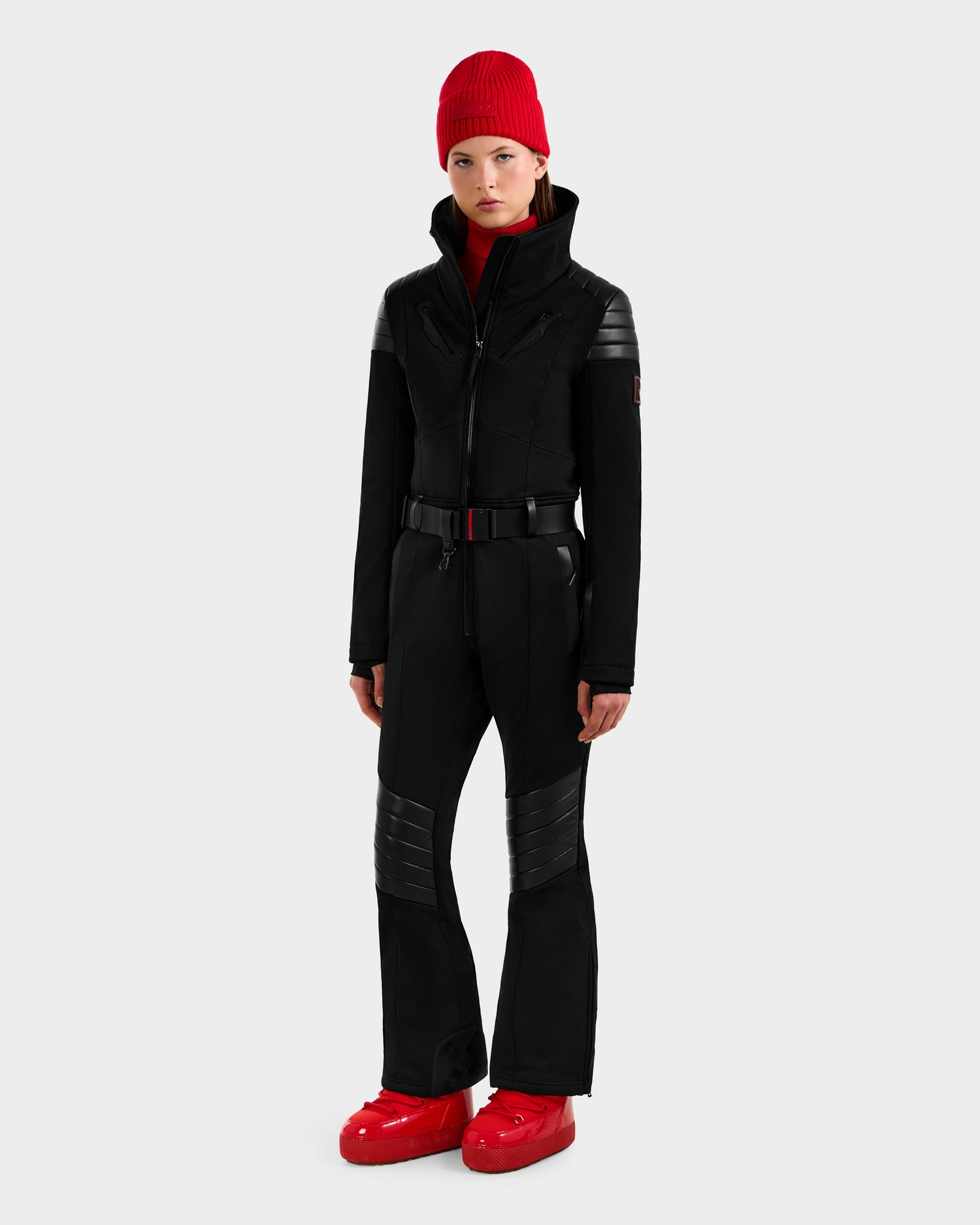 A person models the Rudsak ARLEY - 8124717 Black fitted women's ski suit with quilted details and RECCO rescue tech, styled with a red knit beanie, red turtleneck, and shiny red snow boots against a plain light gray background.