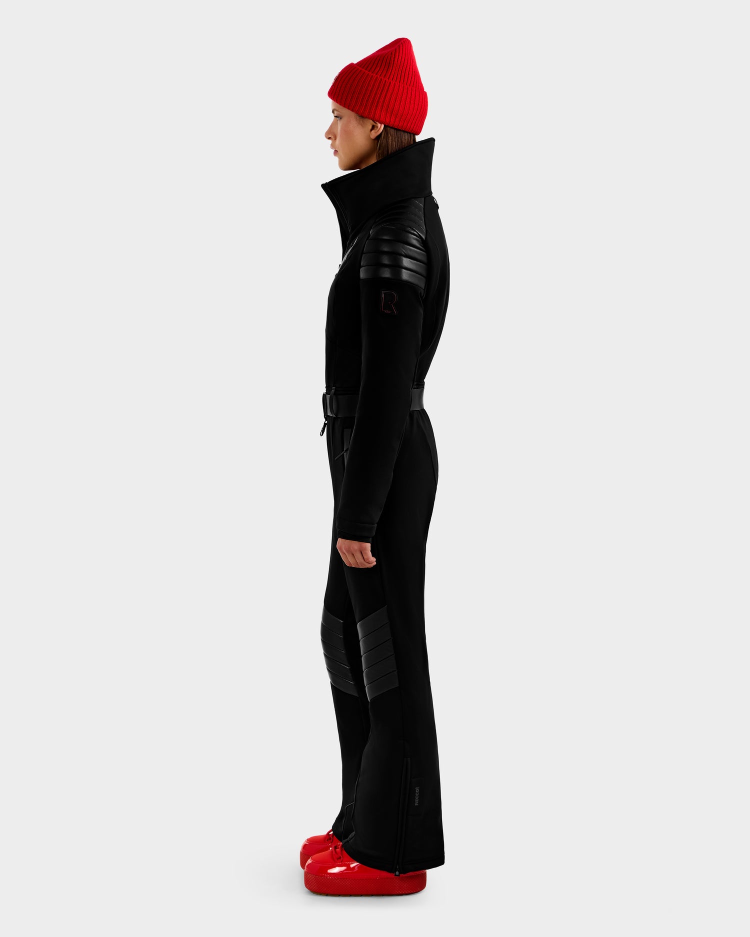 A person in profile wears the Rudsak ARLEY - 8124717 Black fitted ski suit with textured shoulders and knees, paired with a red beanie and shiny red snow boots, standing against a plain light gray background.