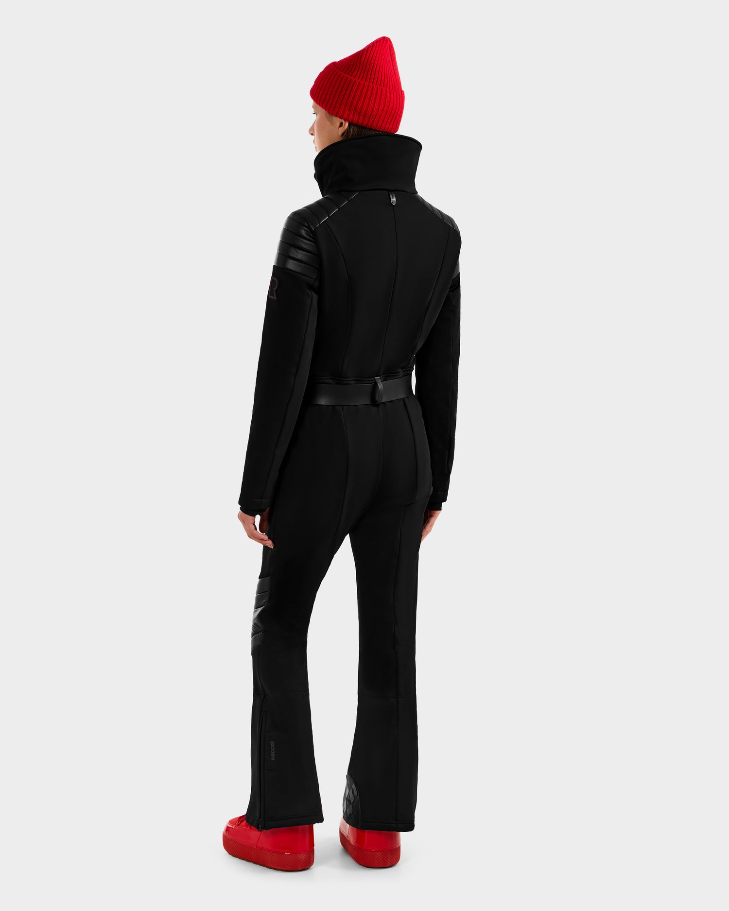 A person faces away, wearing the Rudsak ARLEY - 8124717 Black fitted one-piece ski suit with a high collar, styled with a red beanie and red shoes. The background is plain and light gray.