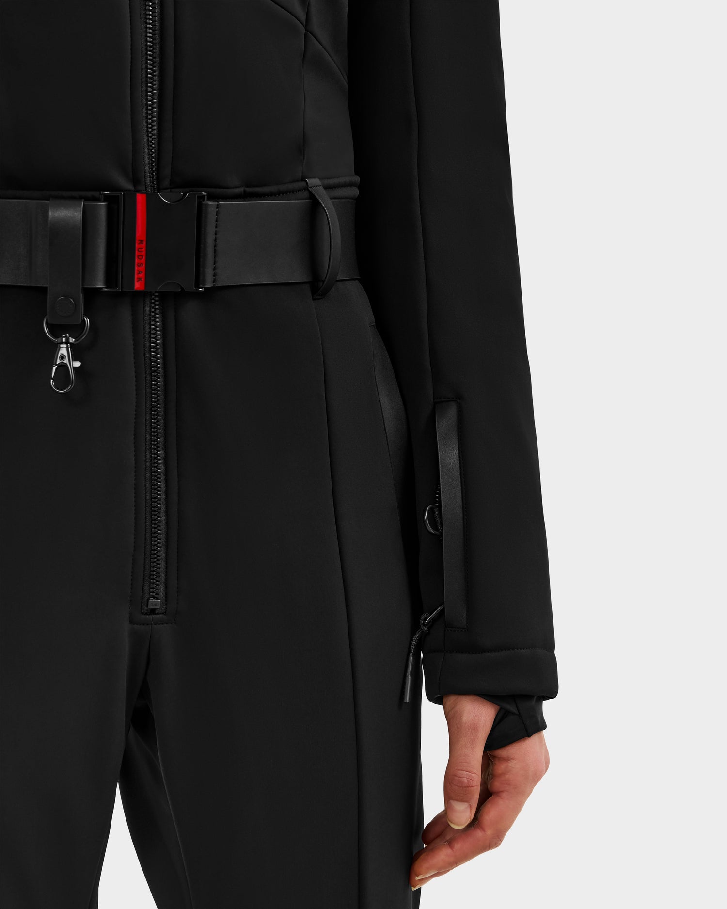 A close-up of a person wearing the ARLEY - 8124717 Black one-piece ski suit by Rudsak, featuring a matching belt with a red accent and sleeve thumb hole—ideal for women’s ski suit enthusiasts.