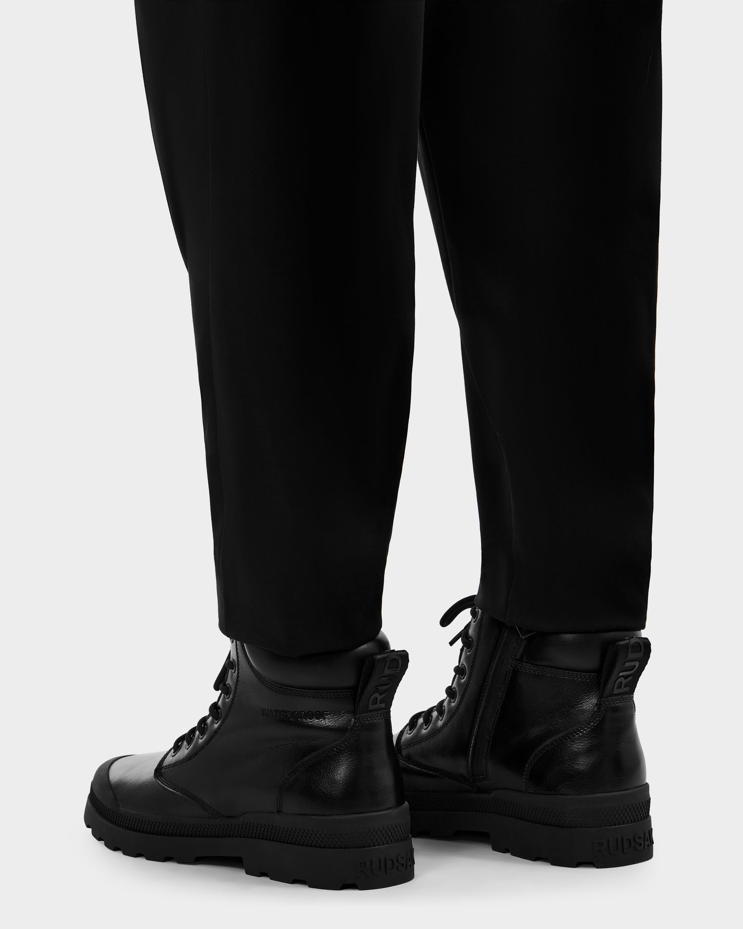 A person wearing black pants and Rudsak ARTUR - M223501 men's boots with side zippers stands on a light gray surface. The ankle boots have rugged soles, weather-treated black leather, and Rudsak branding on the heel.