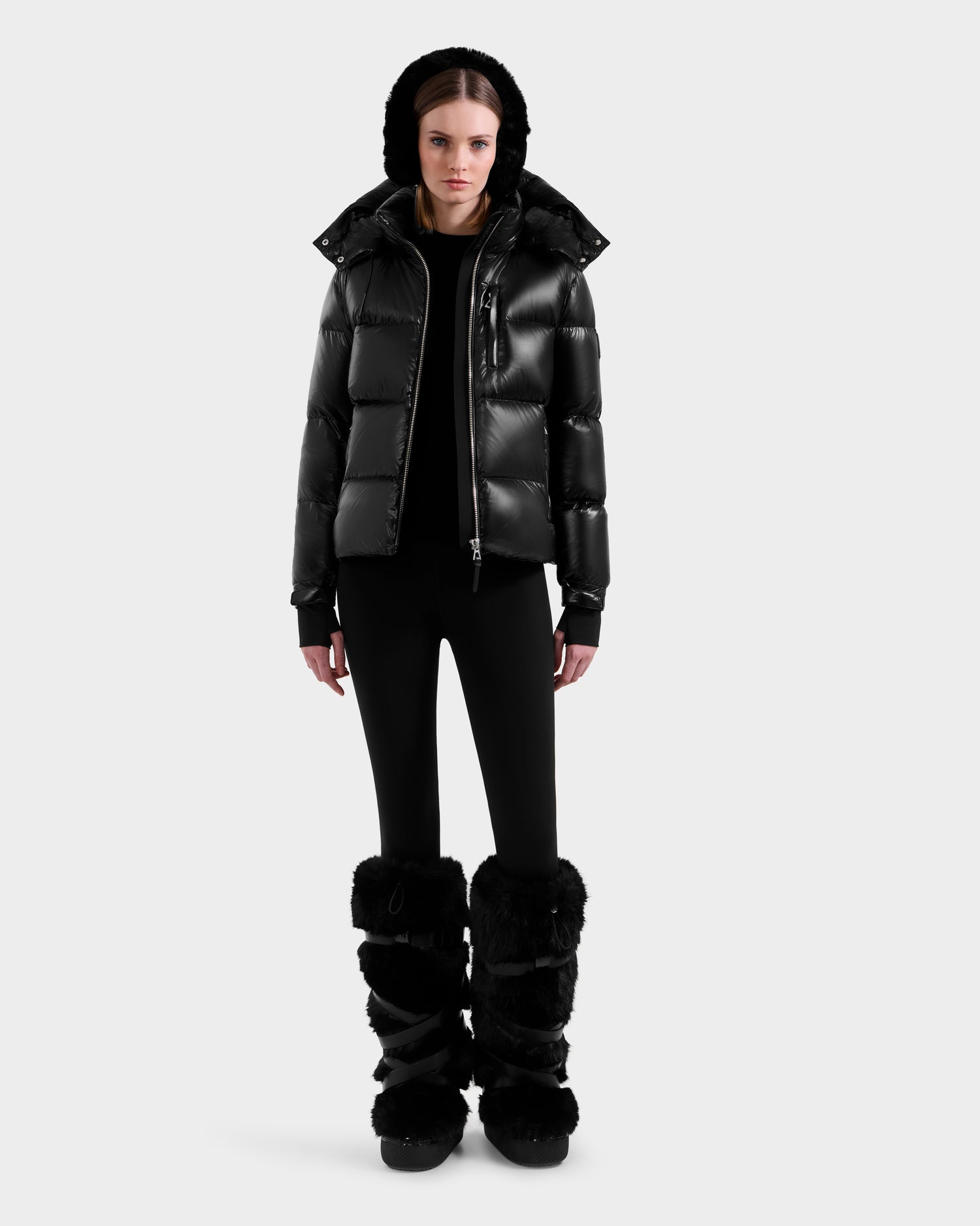 A woman faces forward wearing the Rudsak ASHLYN S - 8124565 Black down puffer jacket with a hood, paired with black leggings and black furry boots, set against a plain light gray background.