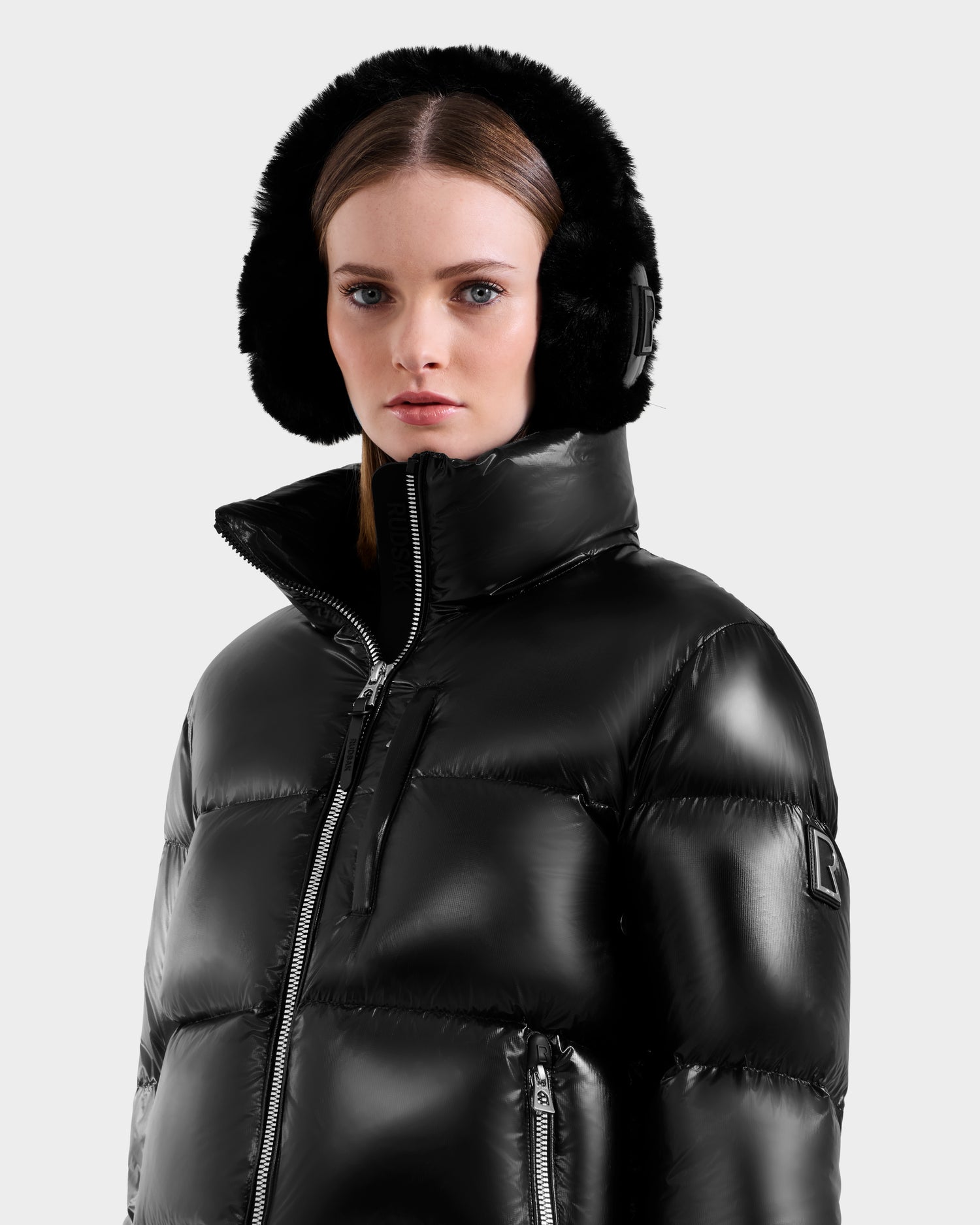 A woman wearing the Rudsak ASHLYN S - 8124565 Black down puffer jacket with a high collar and black fur earmuffs looks at the camera against a plain light background.
