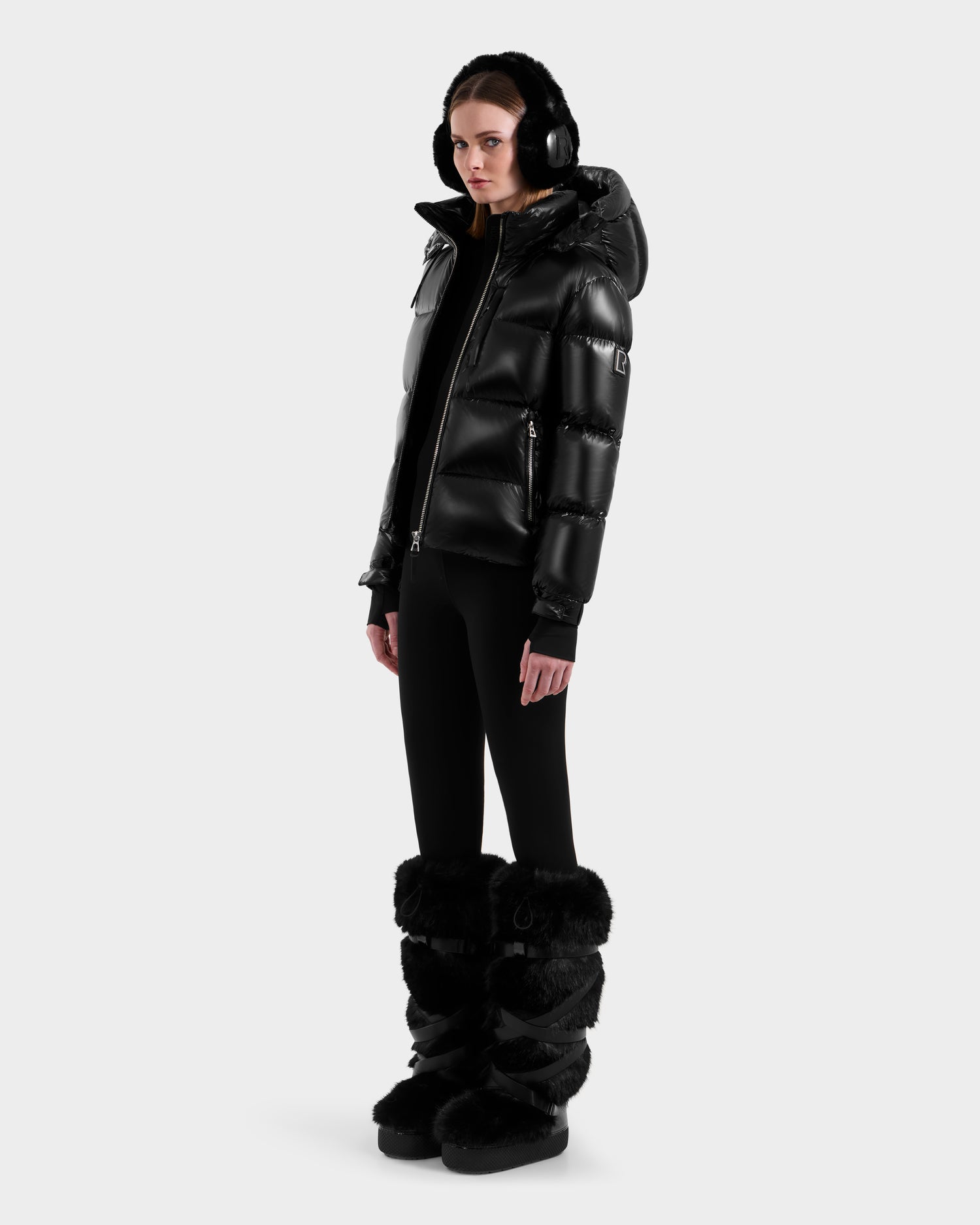 A person wearing the Rudsak ASHLYN S - 8124565 Black women's down puffer jacket with a water-resistant shell, black earmuffs, leggings, and tall fur boots stands against a plain white background.