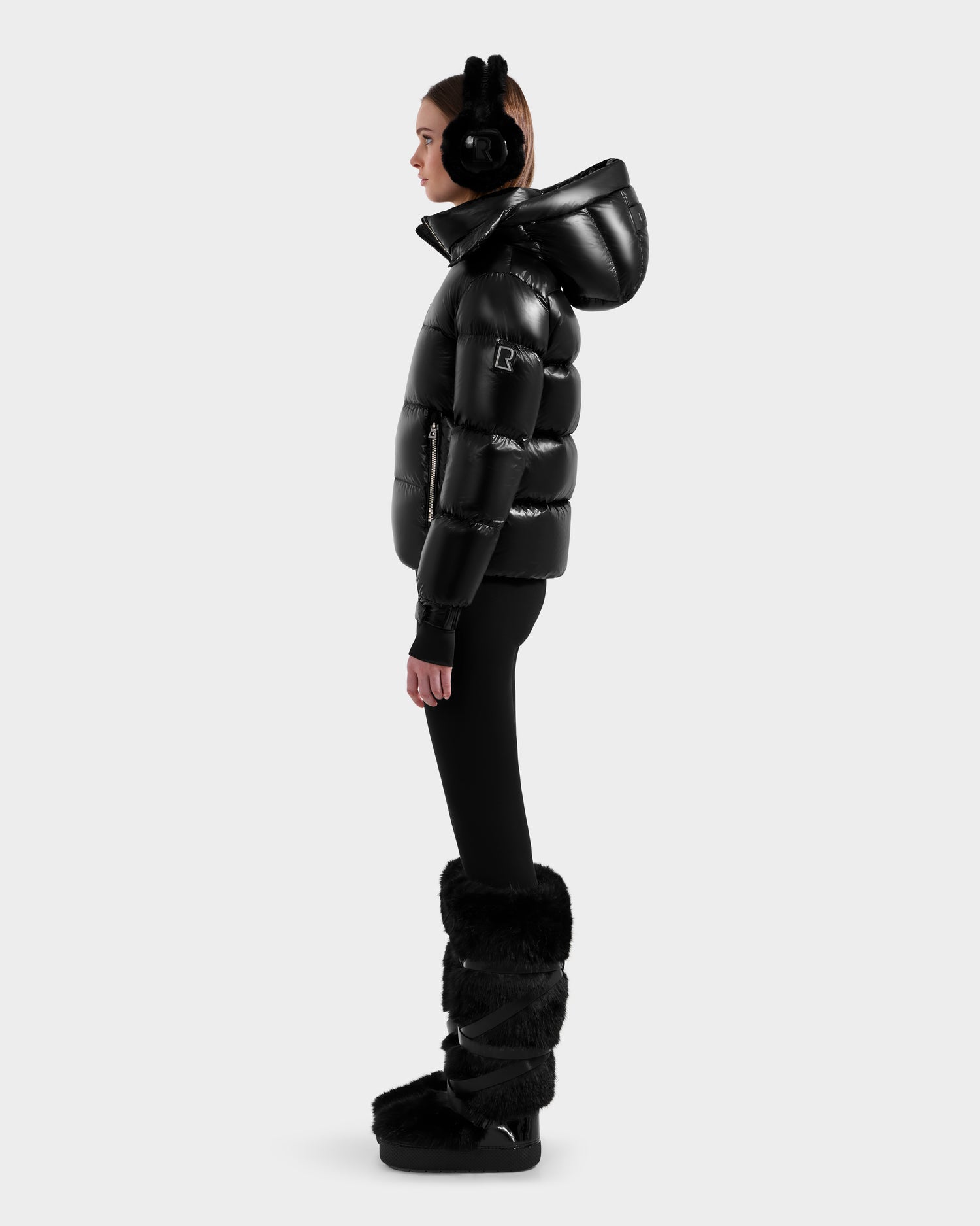 A person in profile wears the Rudsak ASHLYN S - 8124565 Black women’s down puffer with a water-resistant shell and hood, paired with black earmuffs, black leggings, and furry winter boots against a plain light background.
