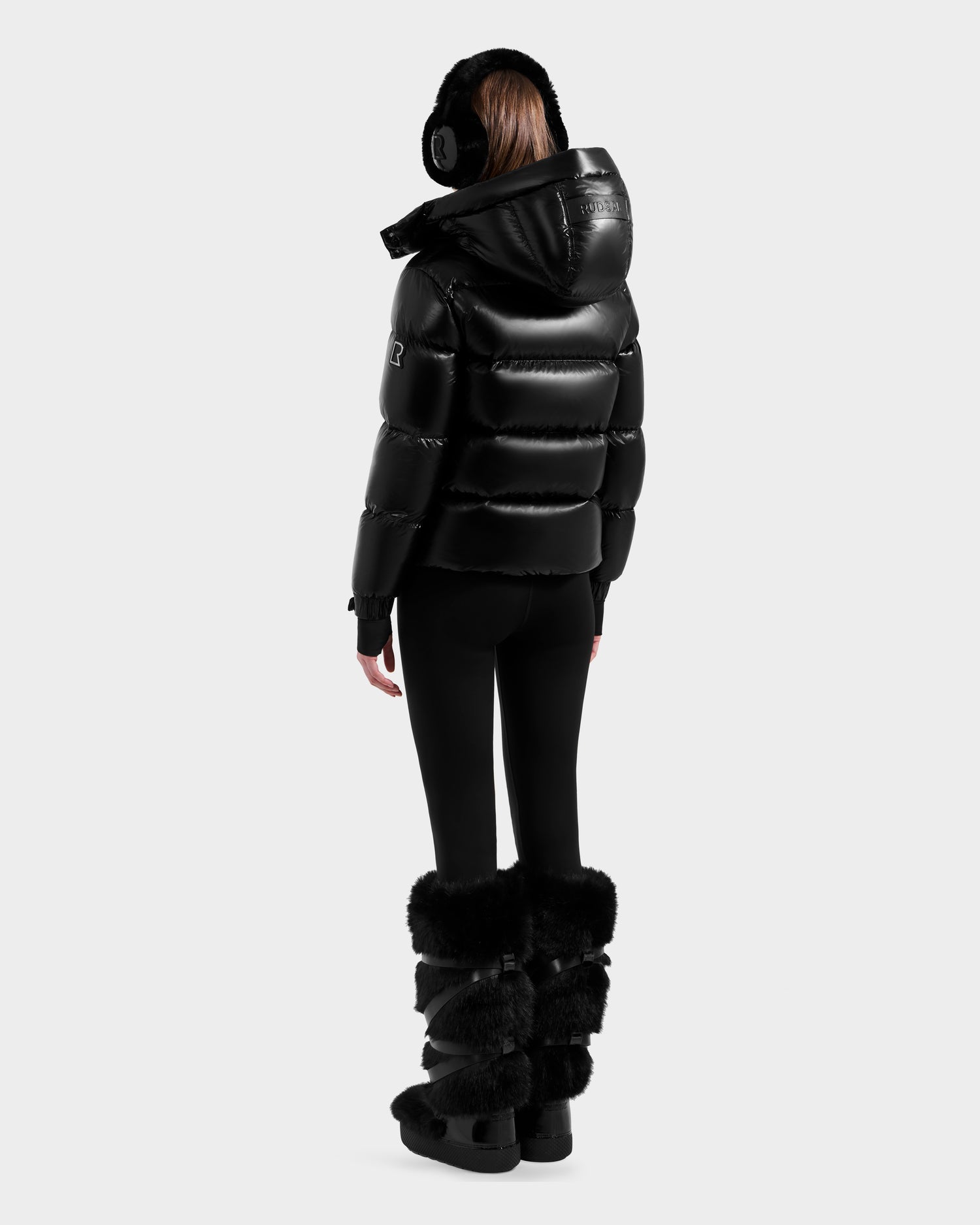 A person stands facing away in a Rudsak ASHLYN S - 8124565 Black women's down puffer jacket with hood, paired with black leggings, fluffy boots, and earmuffs against a light gray background.