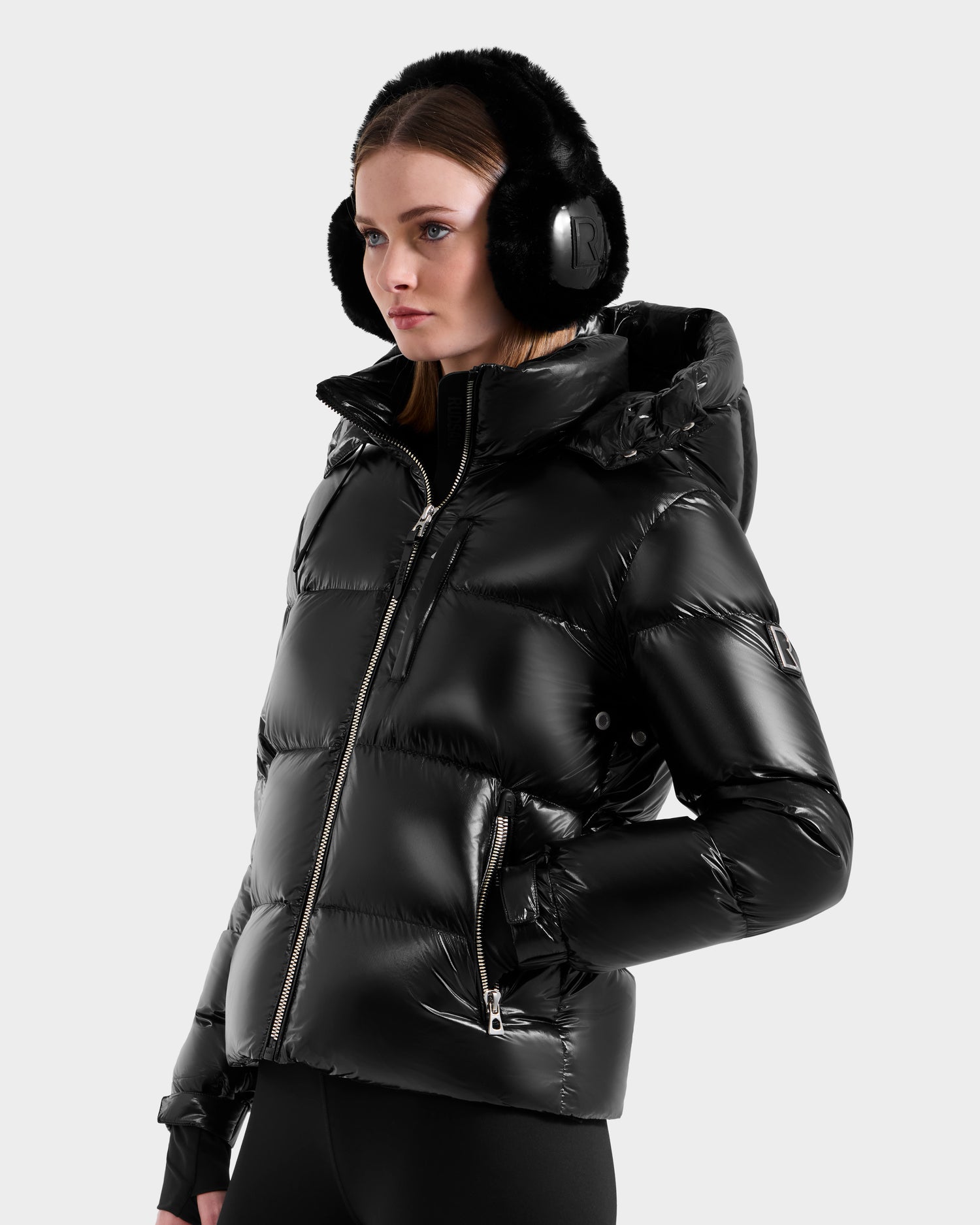 A woman wears the Rudsak ASHLYN S - 8124565 Black down puffer jacket with a hood and zippered pockets, paired with black earmuffs and pants, standing against a plain light gray background.