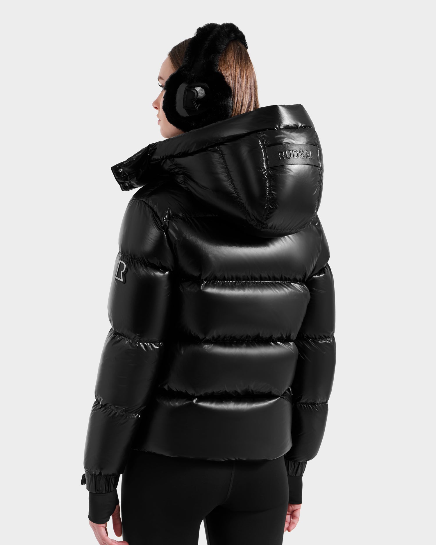 A person in the Rudsak ASHLYN S - 8124565 Black women's down puffer, with RDS-certified down, a logo sleeve patch, shiny water-resistant shell, black earmuffs, and black pants stands facing away on a plain light background.