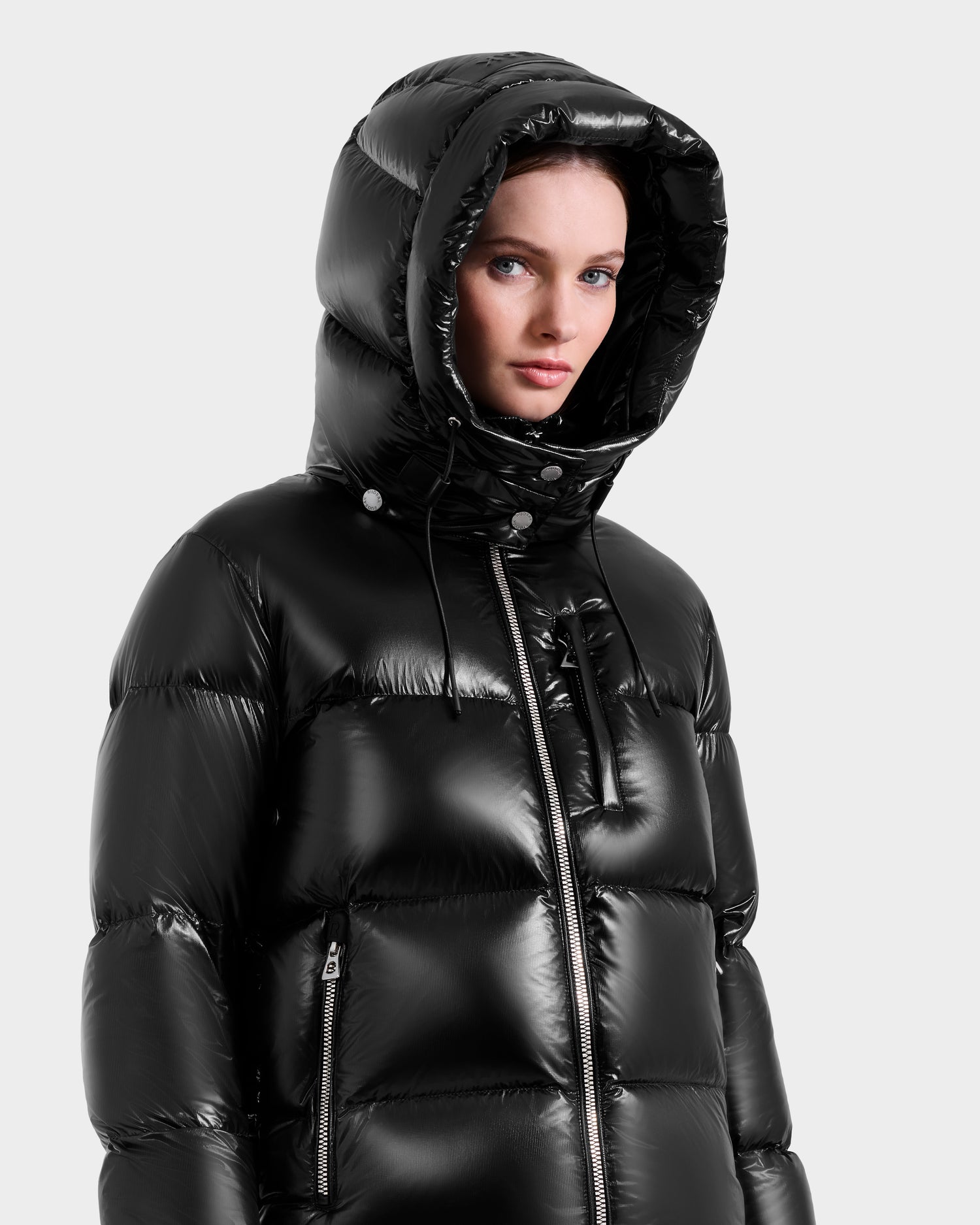A woman wears the Rudsak ASHLYN S - 8124565 Black down puffer with a water-resistant shell and hood up, looking slightly to the side against a plain light gray background.