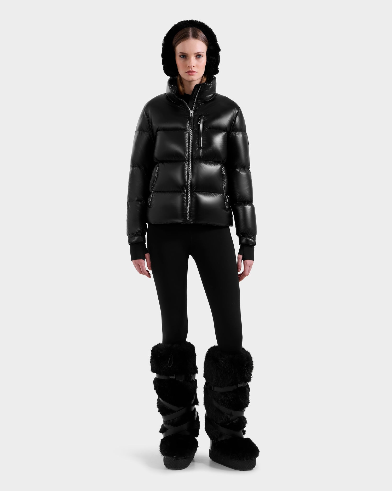 A person faces forward against a plain light background, wearing the Rudsak ASHLYN S - 8124565 Black women’s down puffer jacket with a hood, black leggings, and tall black boots with furry trim.