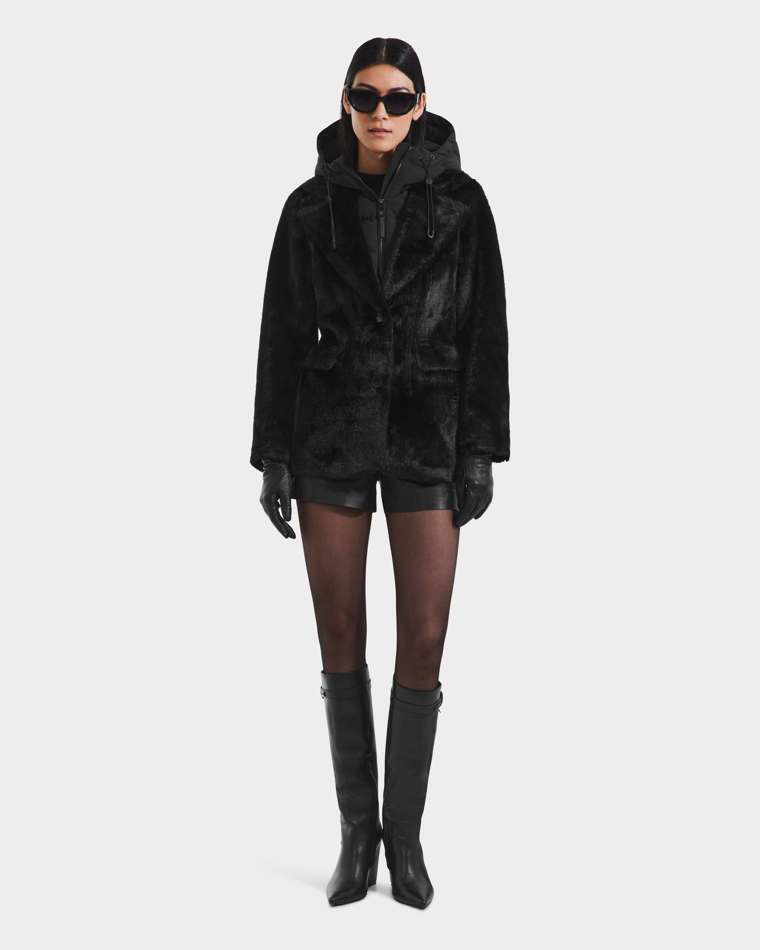 A person models the Rudsak ASPEN F - 8125604 Black jacket—complete with a down-insulated hood, black shorts, tights, knee-high boots, leather gloves, and oversized sunglasses—creating a chic cold-weather look against a white background.