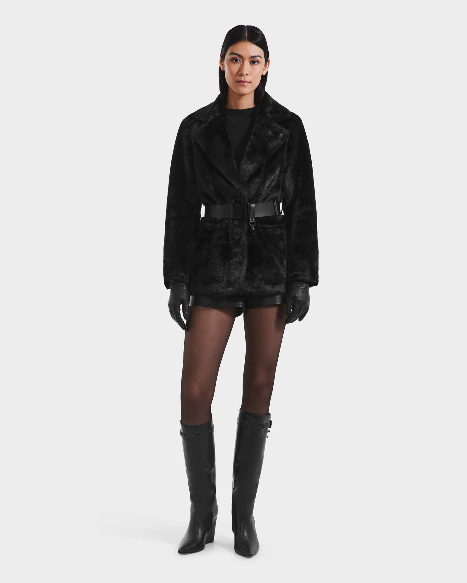 A woman faces forward against a white background, wearing the Rudsak ASPEN F - 8125604 Black women’s faux fur jacket with a belt, paired with black gloves, shorts, sheer tights, and knee-high boots—an elegant winter look.